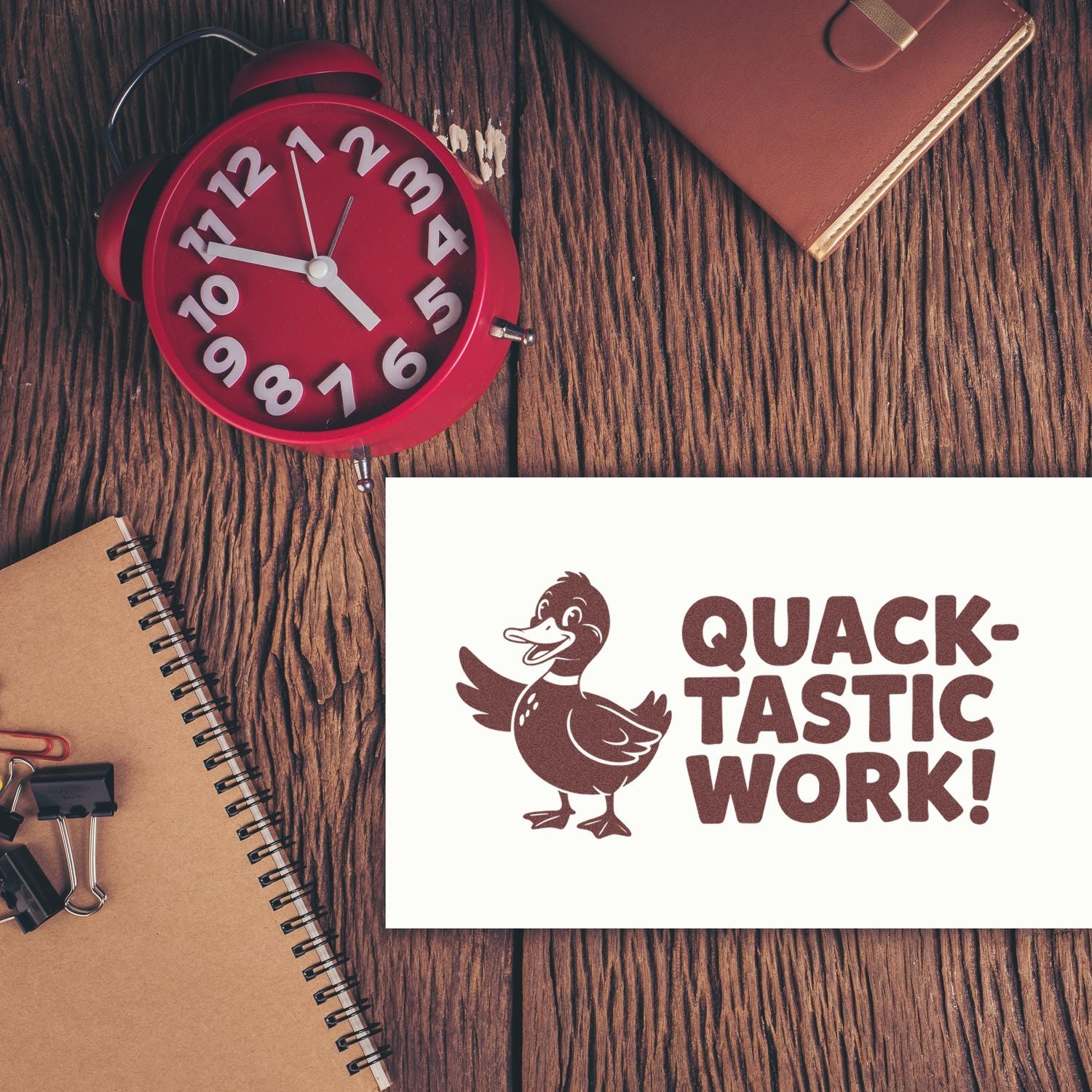 Brown duck stamp seal reading QUACK-TASTIC WORK! on white card, beside a red alarm clock and tan notebook on wood.