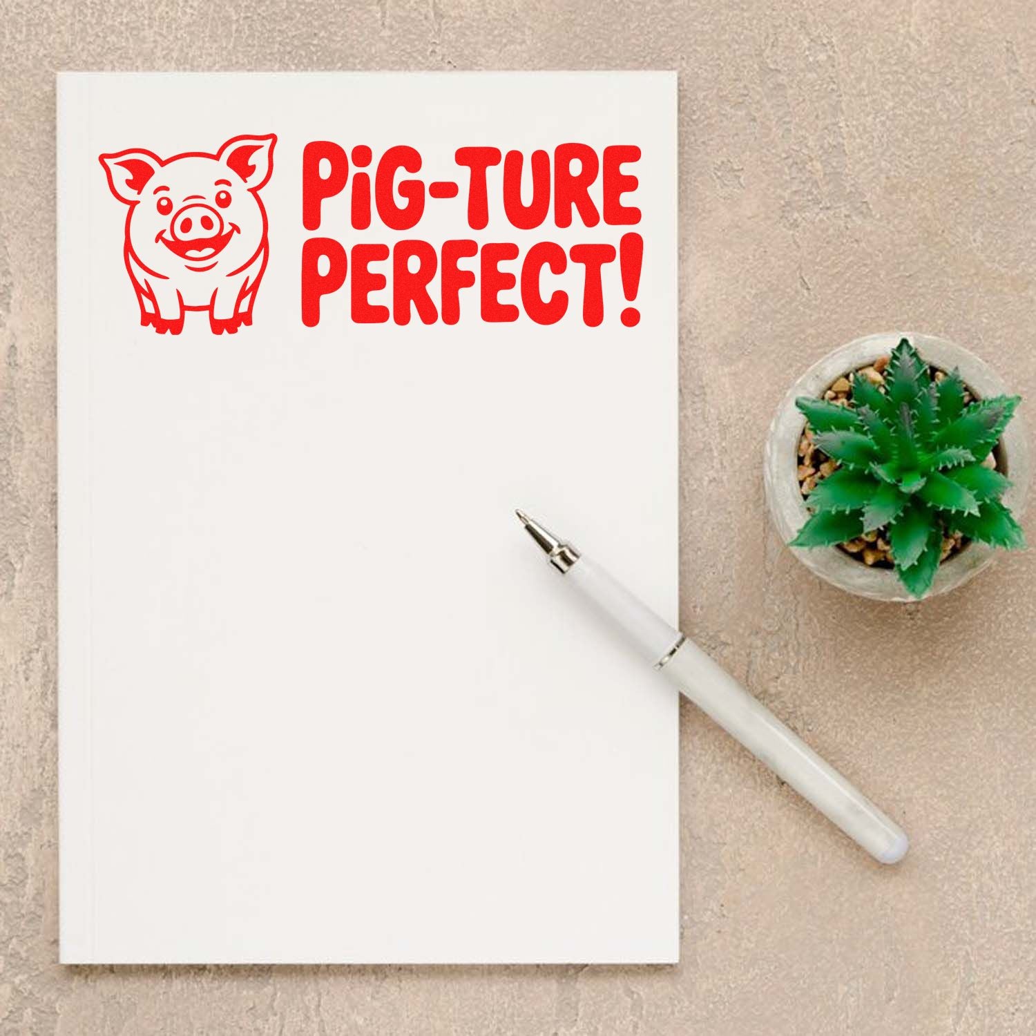 Red pig-shaped stamp seal with PIG-TURE PERFECT! on a white notebook, shown with a white pen on a beige background.