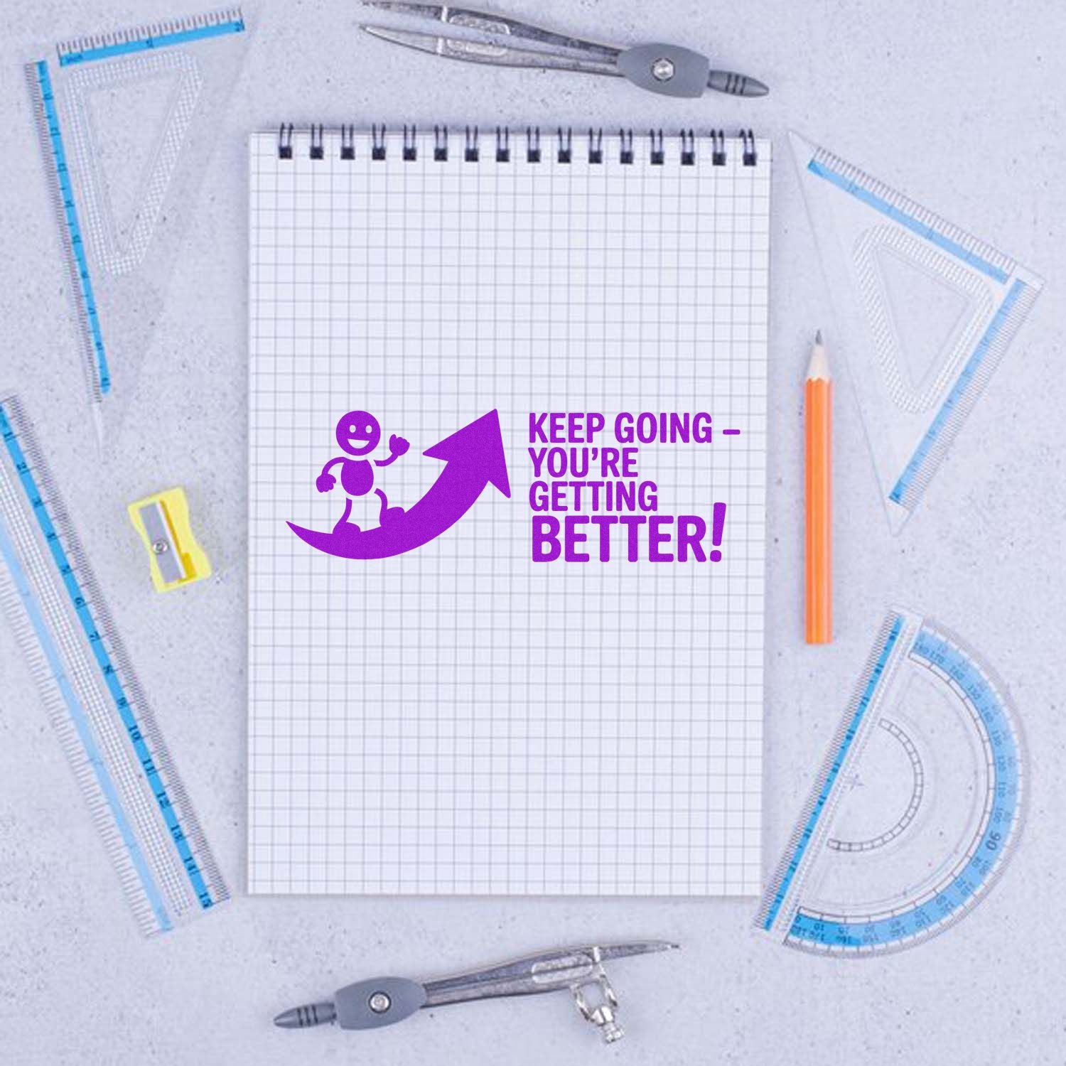 Purple motivational stamp seal on graph notebook: Keep goingyoure getting better! with arrow, surrounded by drafting tools.