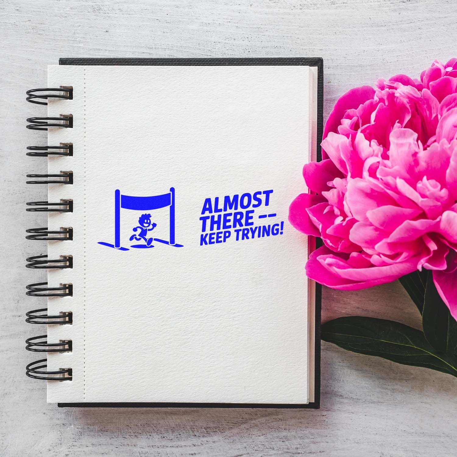 Blue motivational stamp seal reading Almost therekeep trying! on a white spiral notebook next to bright pink flowers.