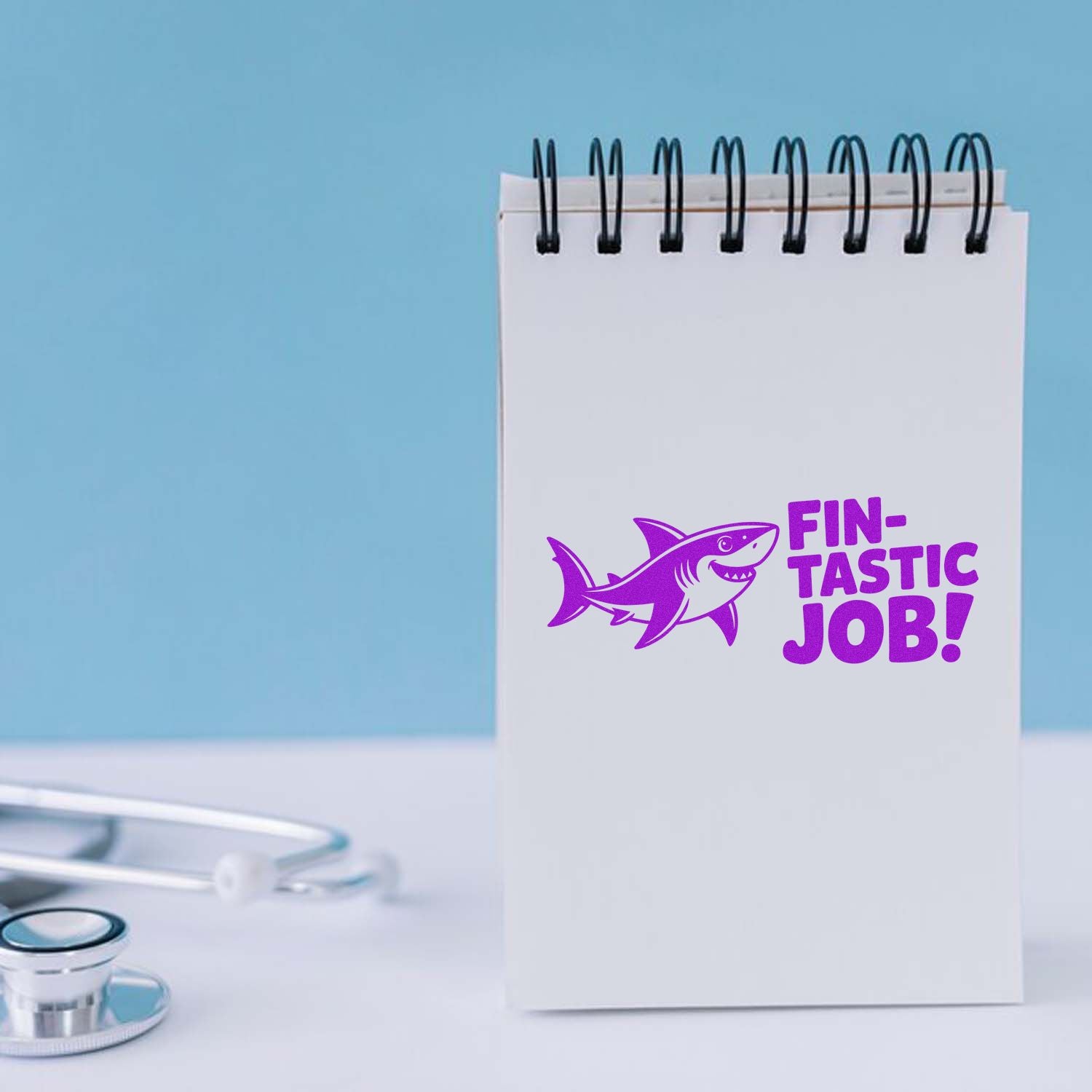 Purple custom rubber stamp on white spiral notepad reads Fin-tastic Job! with shark graphic; blue background and stethoscope.