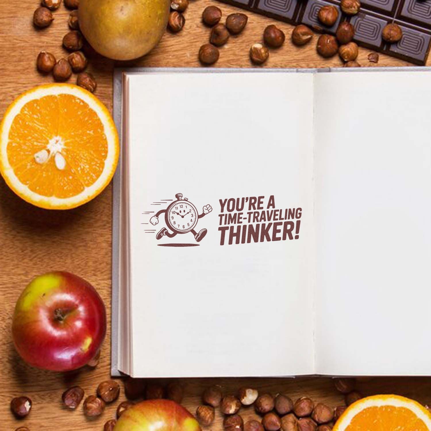 Brown stamp seal on open white notebook reading Youre a time-traveling thinker! with clock graphic, on wood desk with oranges and apples.
