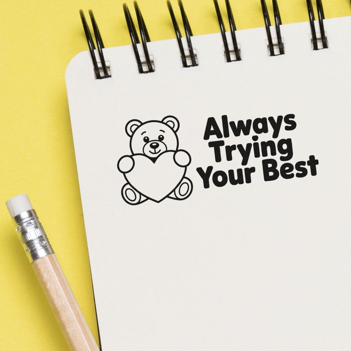 Black teddy bear heart stamp seal with Always Trying Your Best on a white spiral notebook, yellow background, and pencil.