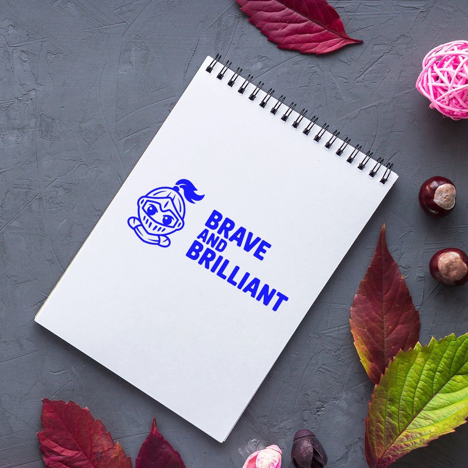 Blue Brave and Brilliant stamp with ninja mascot on white spiral notebook, framed by red and green autumn leaves on gray textured background