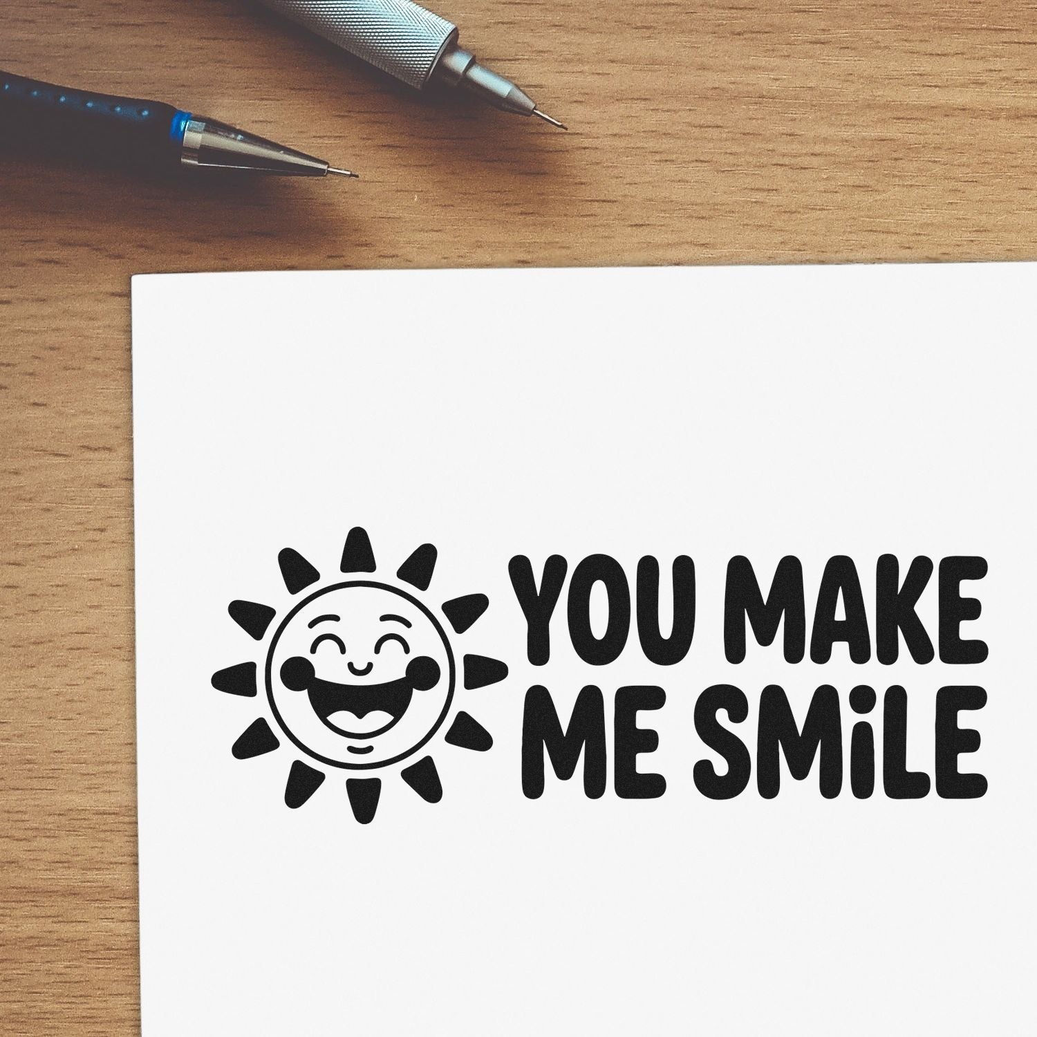 Black smiley sun rubber stamp and seal reading YOU MAKE ME SMILE on white paper, with pens on a wood desk.