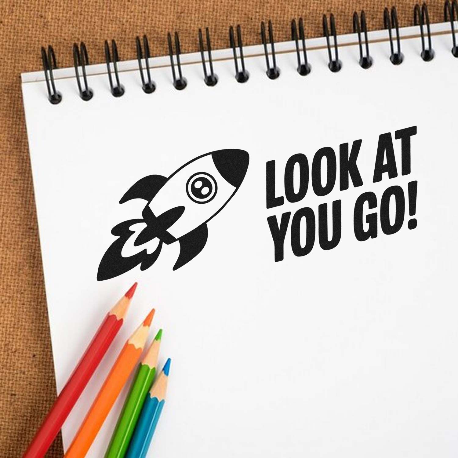 Black rocket stamp and seal with LOOK AT YOU GO! on a white spiral notebook, accented by red, orange, green, and blue pencils.
