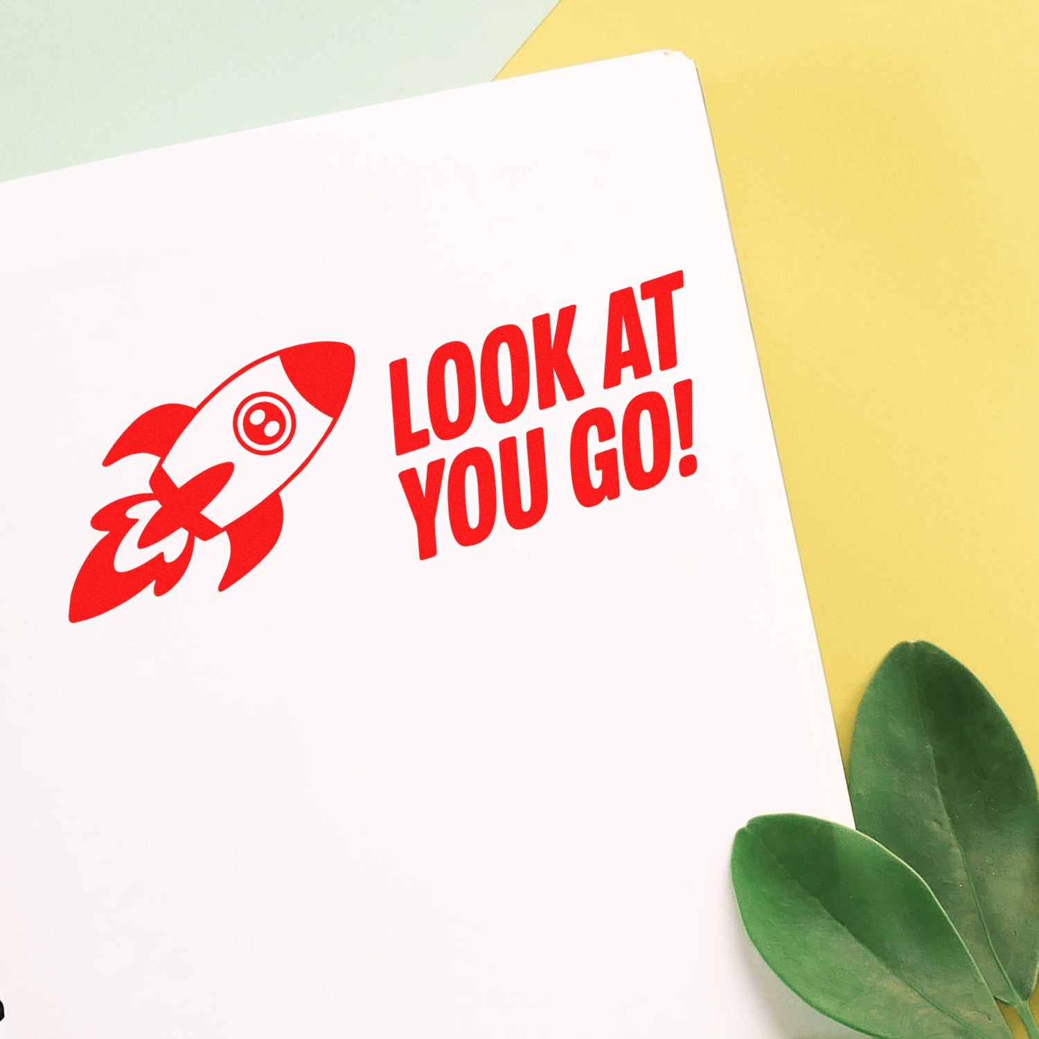 Red rocket stamp seal with LOOK AT YOU GO! on white paper, against a pastel yellow background and green leaves.