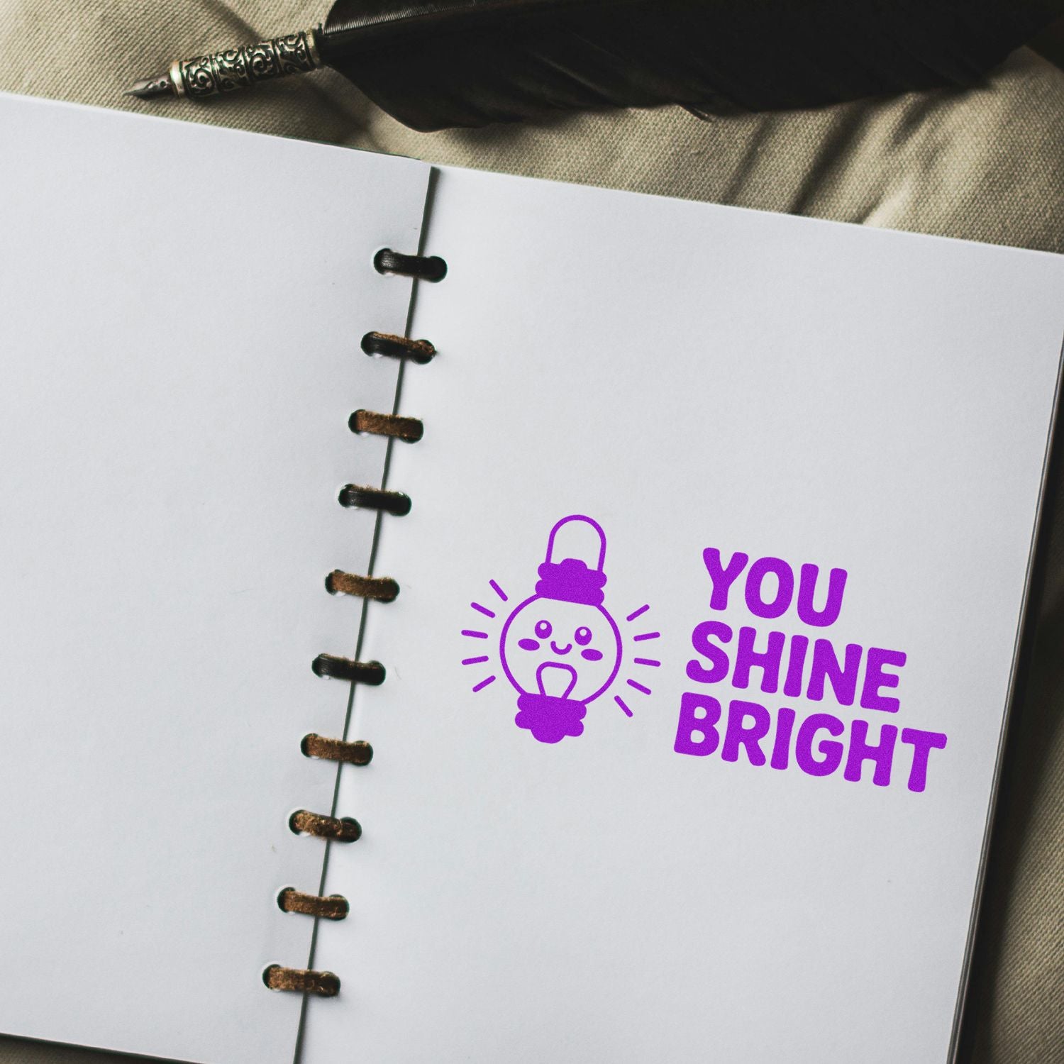 Purple You Shine Bright stamp and lightbulb seal on white notebook pages, with a fountain pen and black lacing.