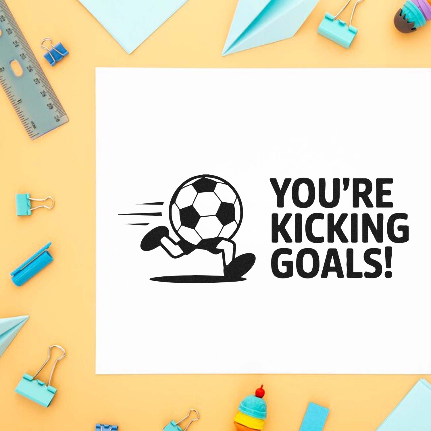 Soccer stamp and seal design in black on white paper with running ball icon and Youre kicking goals! on a yellow desk with blue stationery.