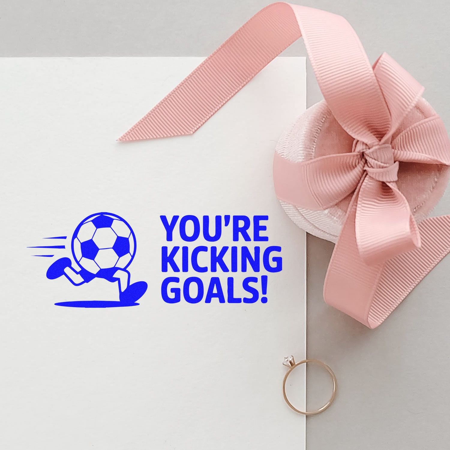 Blue soccer stamp saying "YOU'RE KICKING GOALS!" on white card, topped with a pink ribbon seal and gold ring.