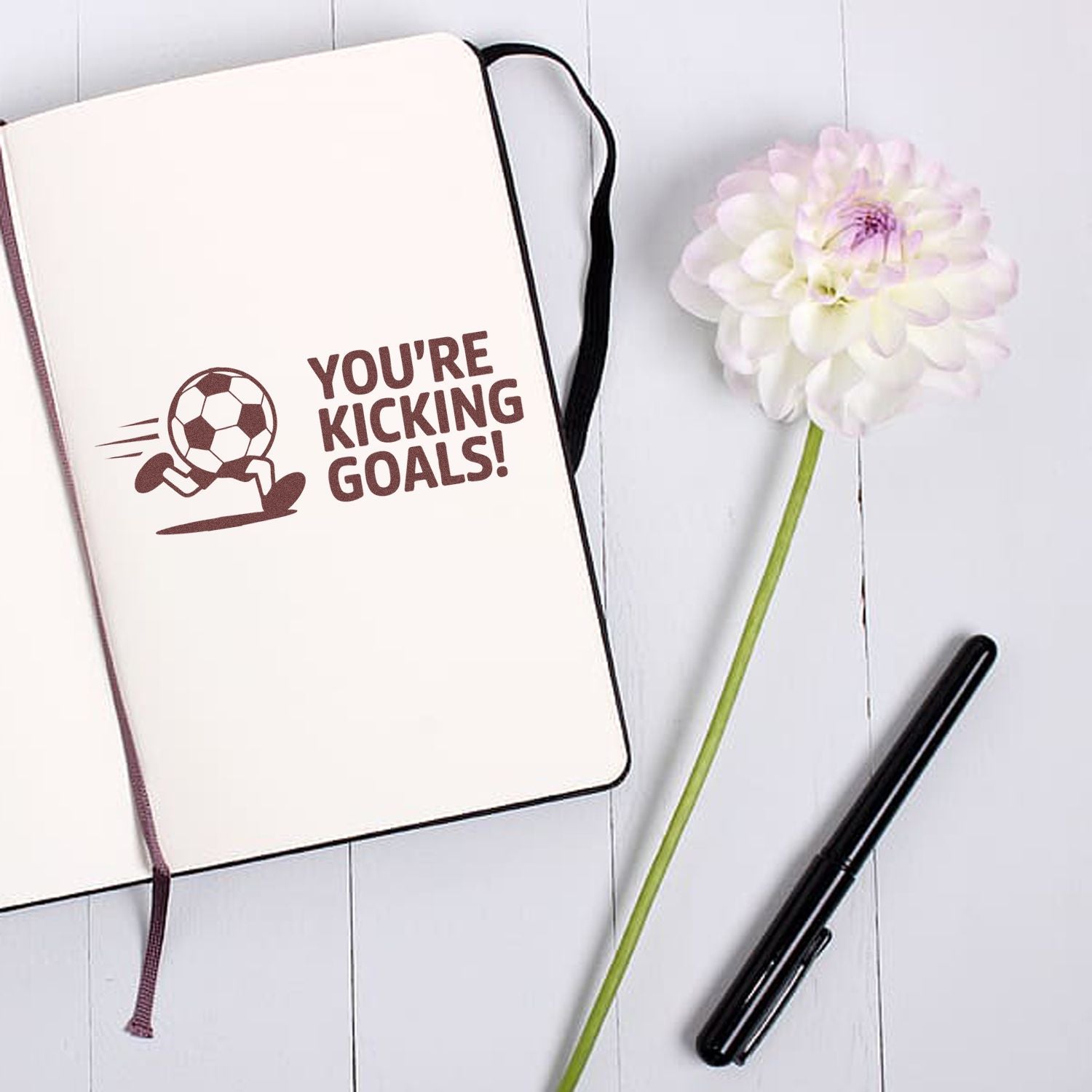 Brown soccer stamp and seal on cream notebook with Youre kicking goals!, white flower, and black pen.
