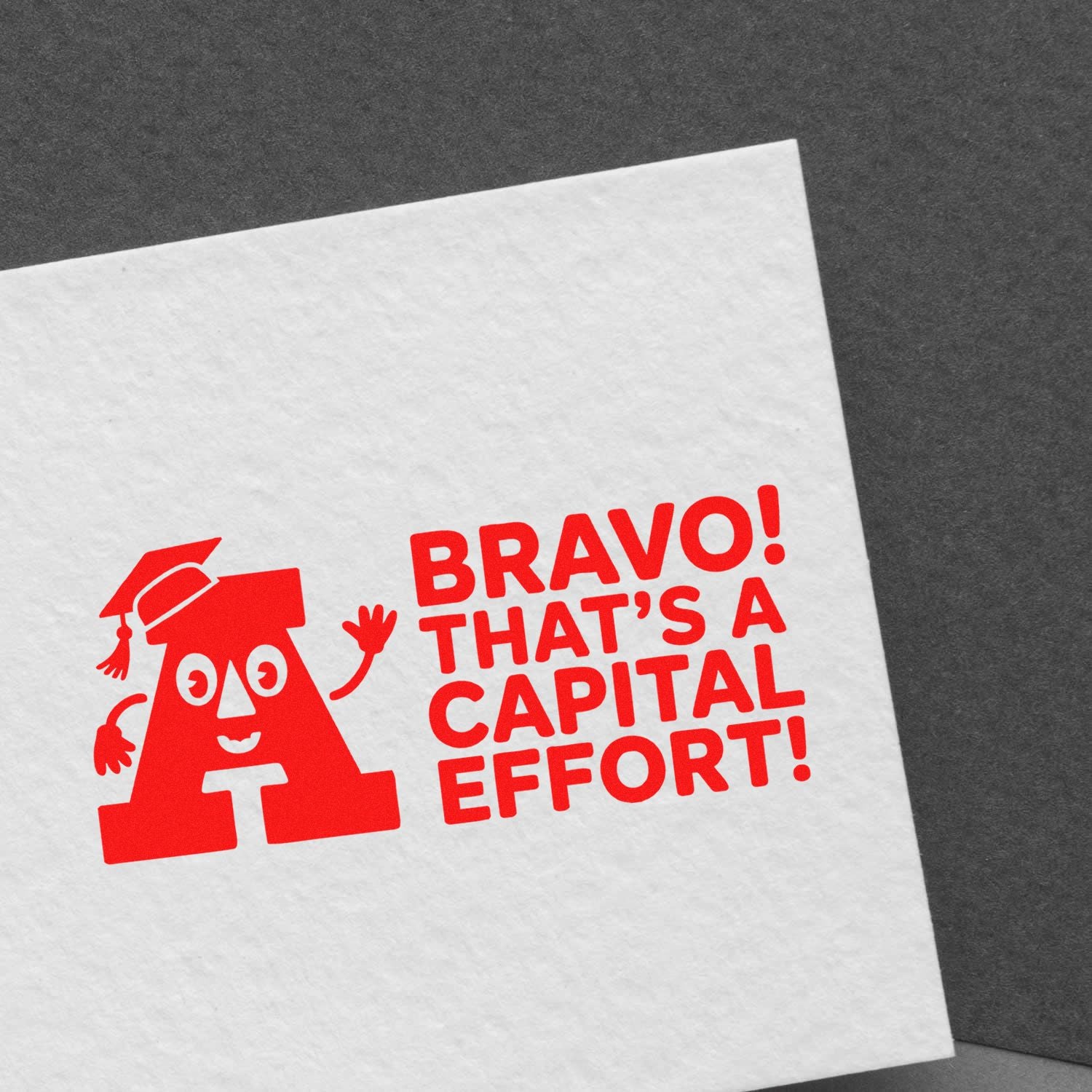 Red stamp seal on white paper reading BRAVO! THATS A CAPITAL EFFORT! with a smiling capital A character.