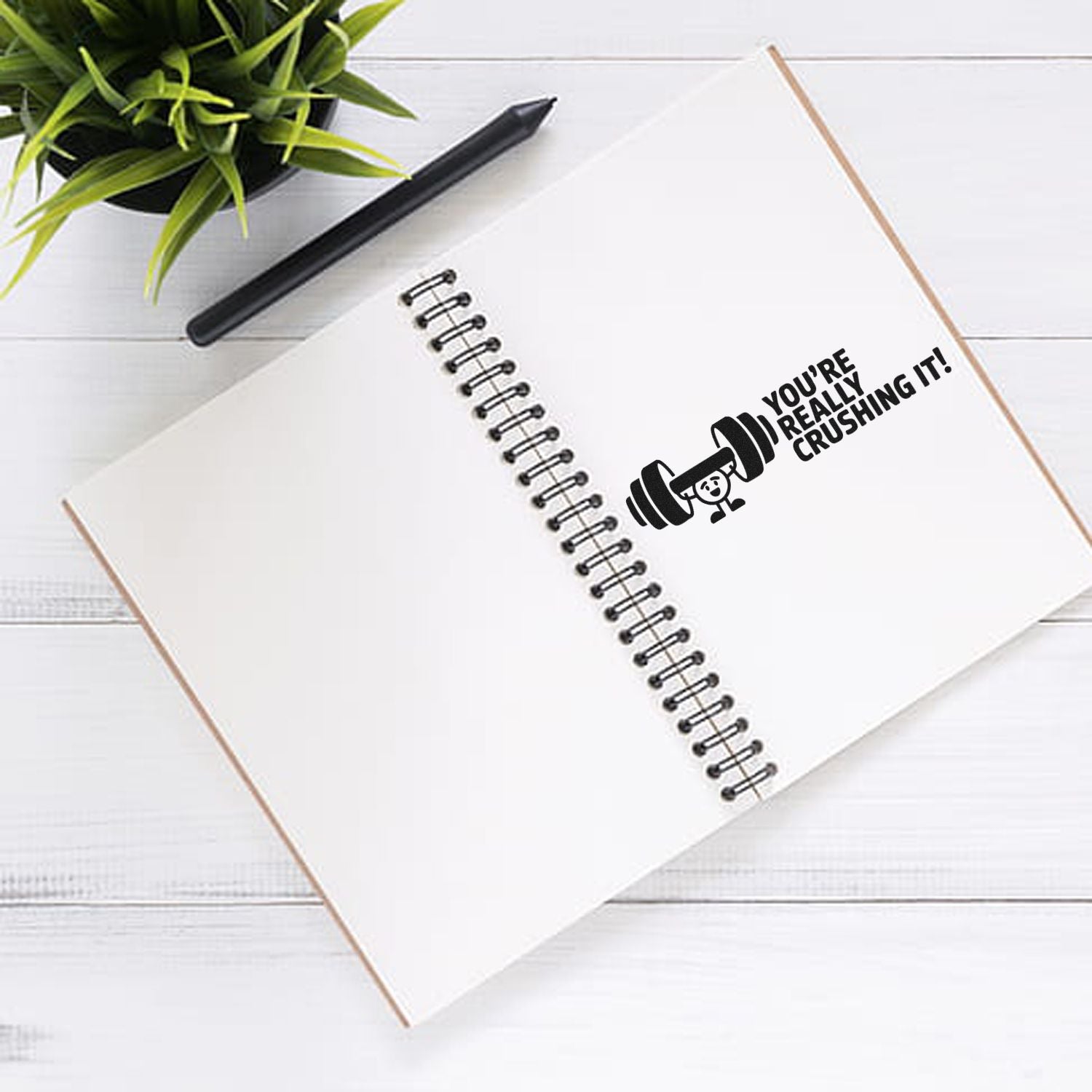 Black motivational stamp seal on a white spiral notebook reading Youre really crushing it! with dumbbell icon, pen, and green plant.