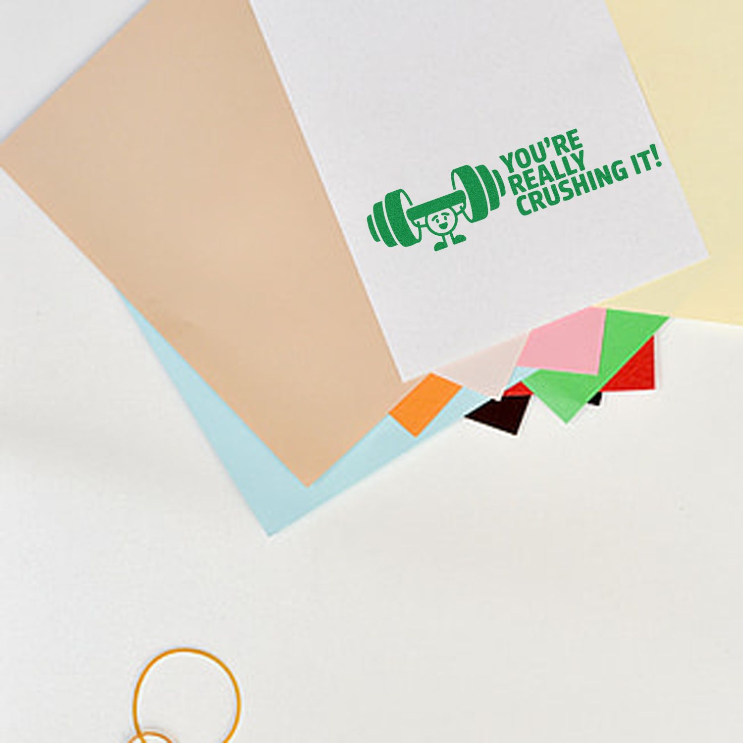 Green motivational stamp seal with dumbbell and Youre really crushing it! on white paper, over pastel stationery sheets.