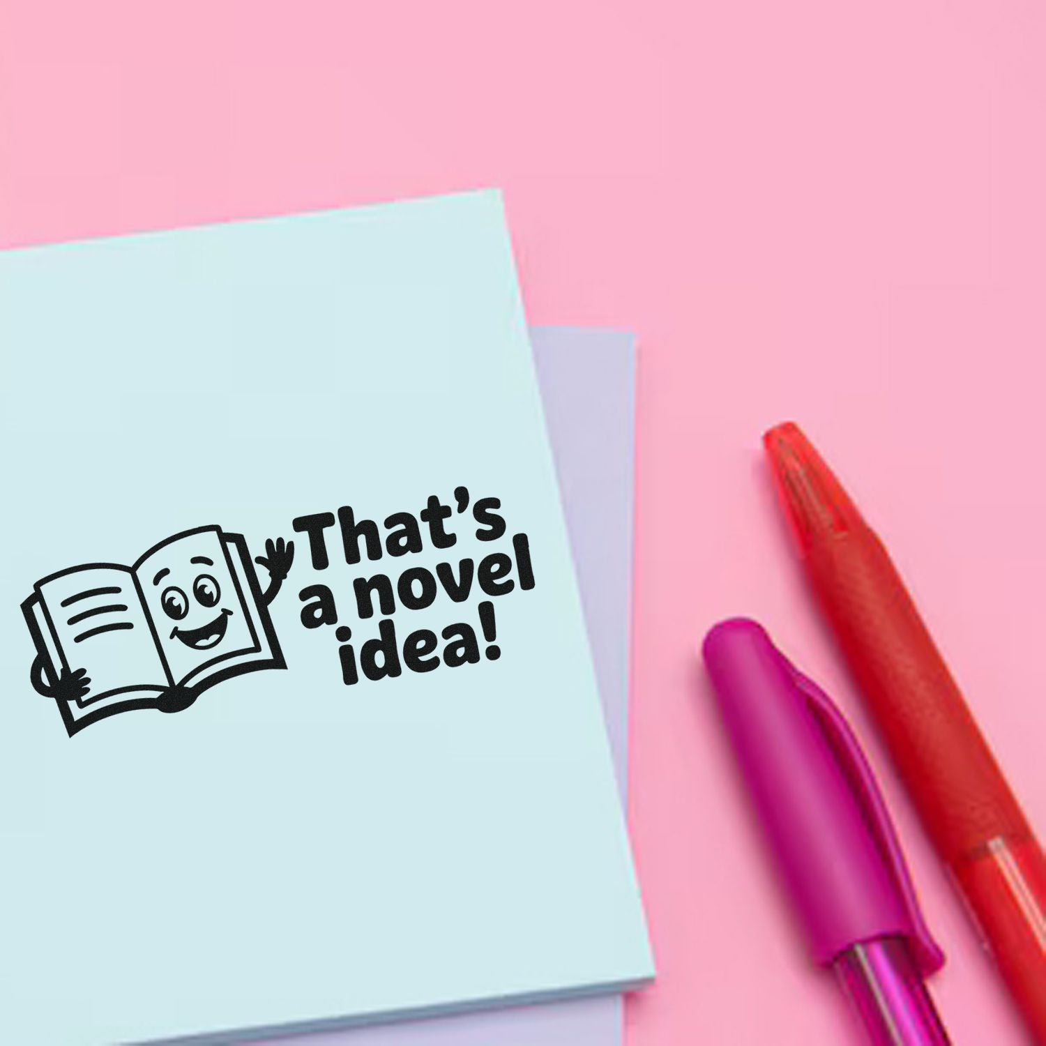 Book-themed rubber stamp seal on a light blue card reading Thats a novel idea! with red and purple pens on pink.