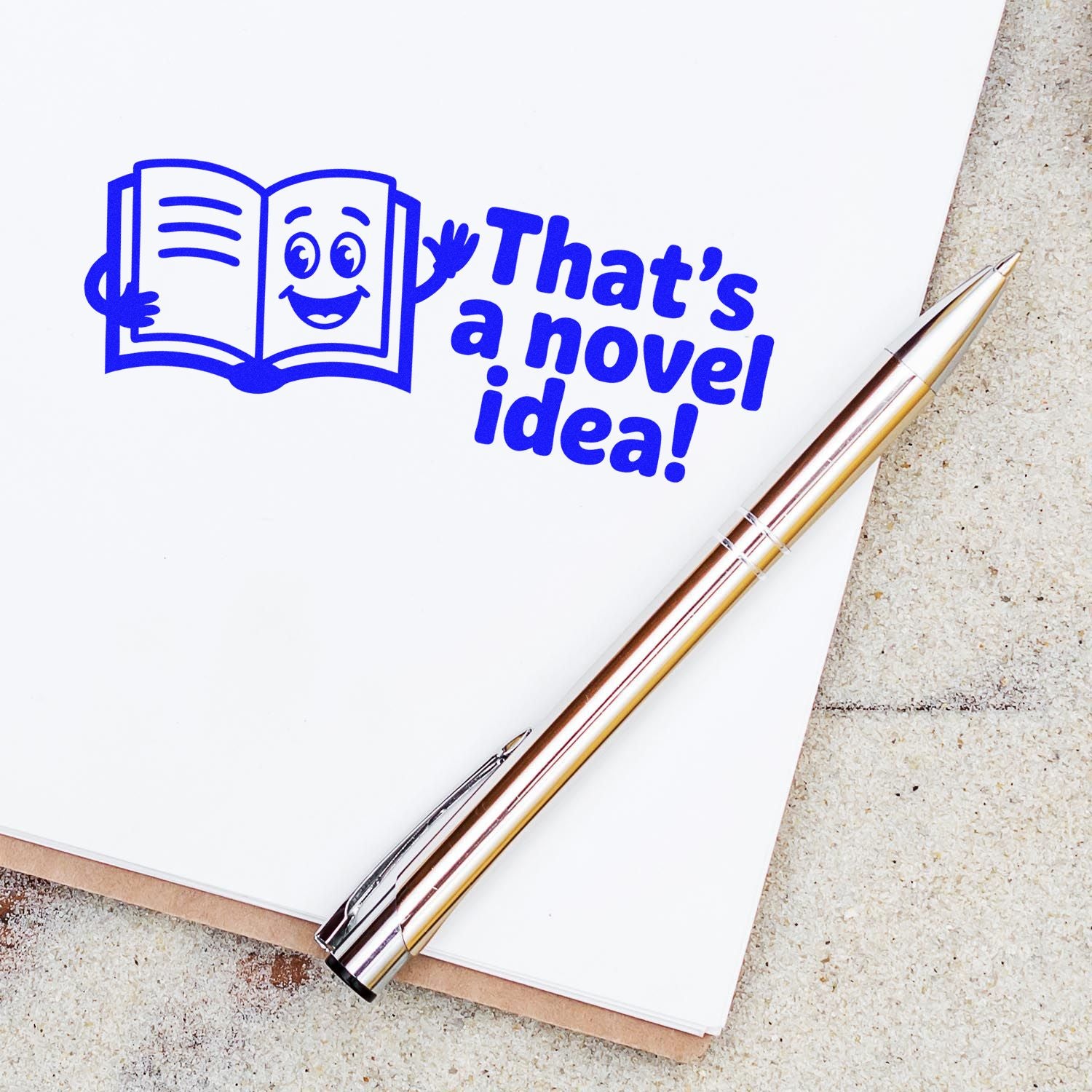 Blue custom stamp with smiling book and Thats a novel idea! in bold blue, beside a gold pen on white paper.