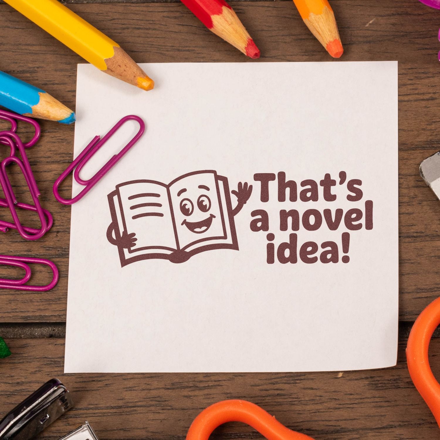 Brown novelty stamp reading Thats a novel idea! with a smiling book on white paper, surrounded by colorful pencils and magenta clips.