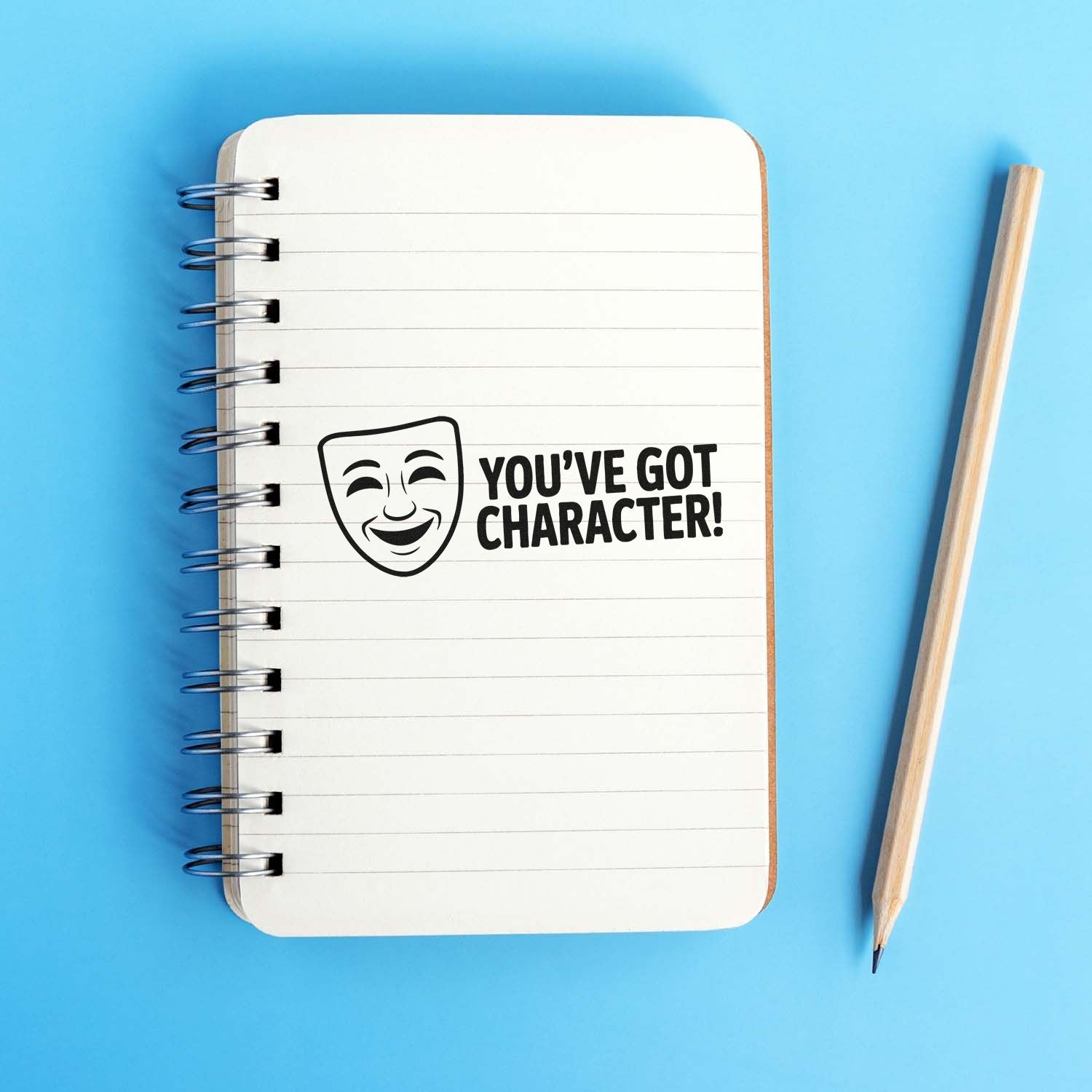 Black stamp seal with theater mask and YOUVE GOT CHARACTER! on a white spiral notebook, blue background, pencil beside it.