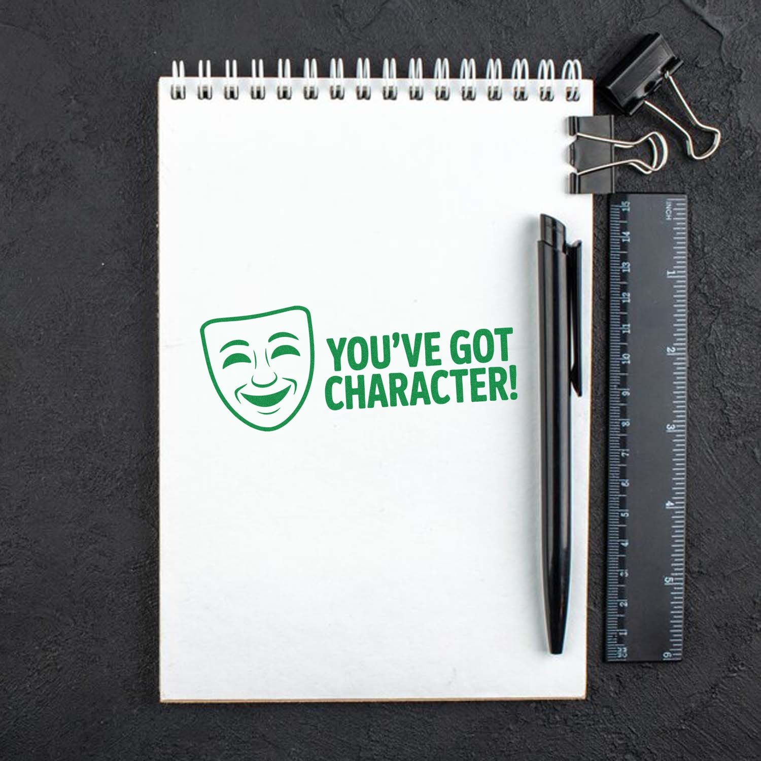 Green theater mask stamp and seal reading Youve Got Character! on white spiral notebook with black pen and ruler.