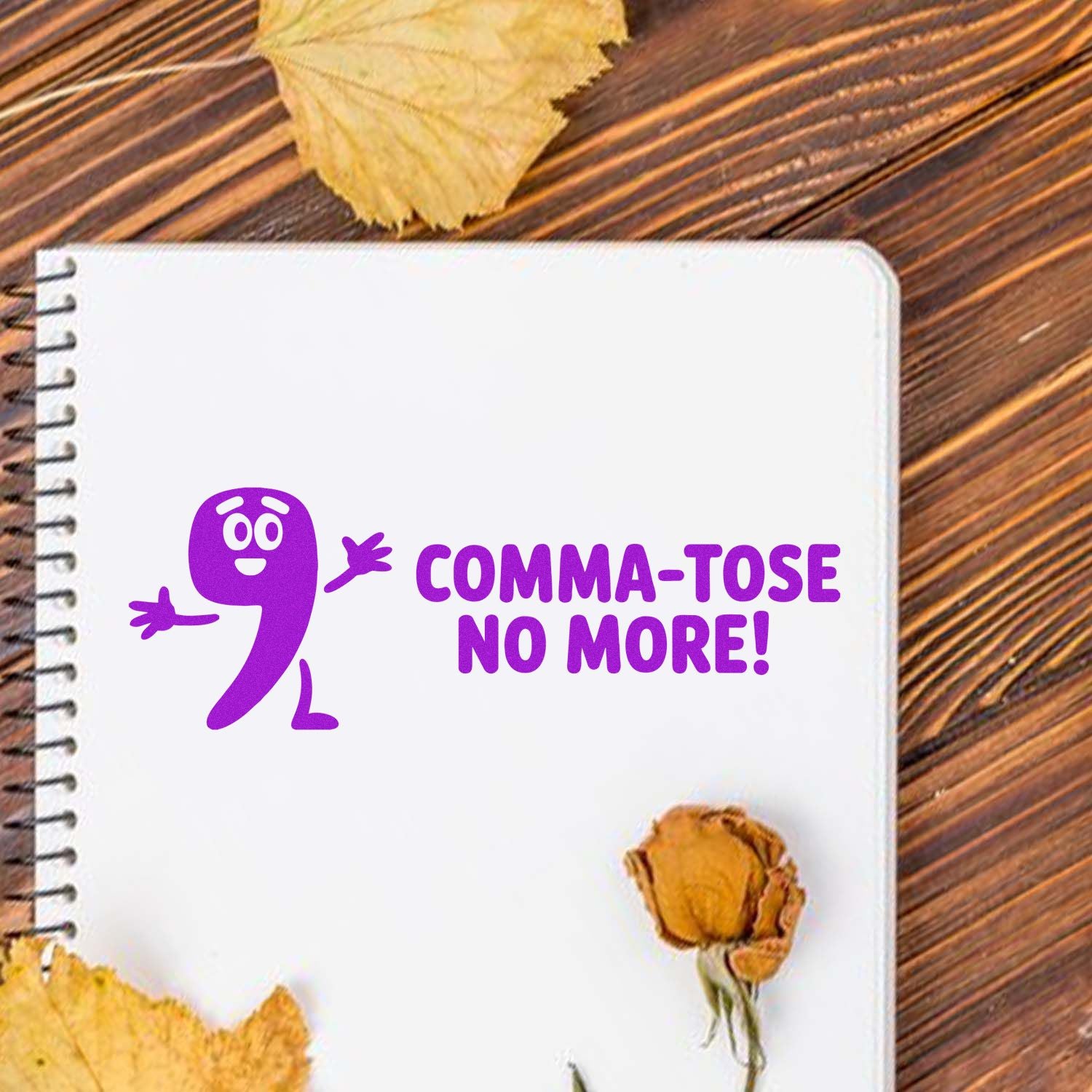 Purple stamp seal with comma mascot and Comma-tose No More! on white notebook, with dried leaves on wood.