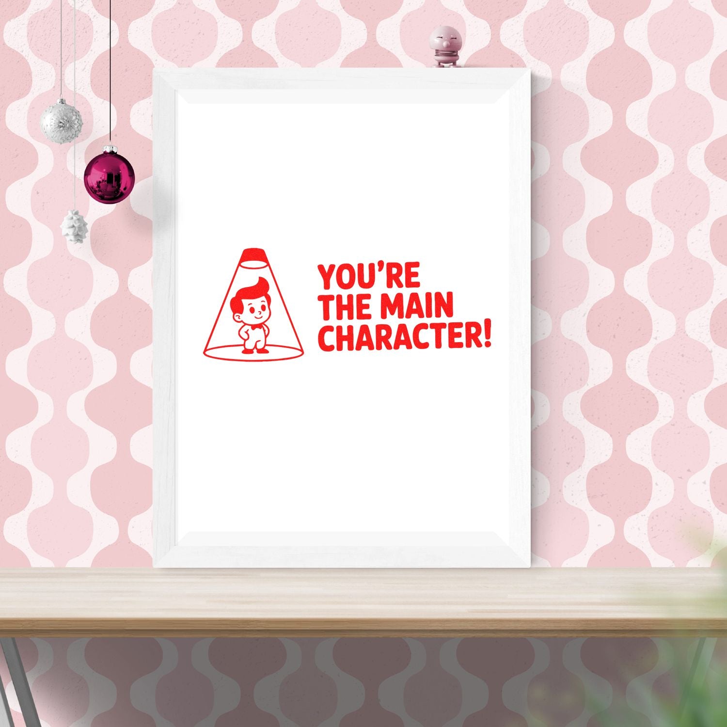 Red stamp seal with cartoon character and Youre the Main Character! on a white framed poster against a pink patterned wall.