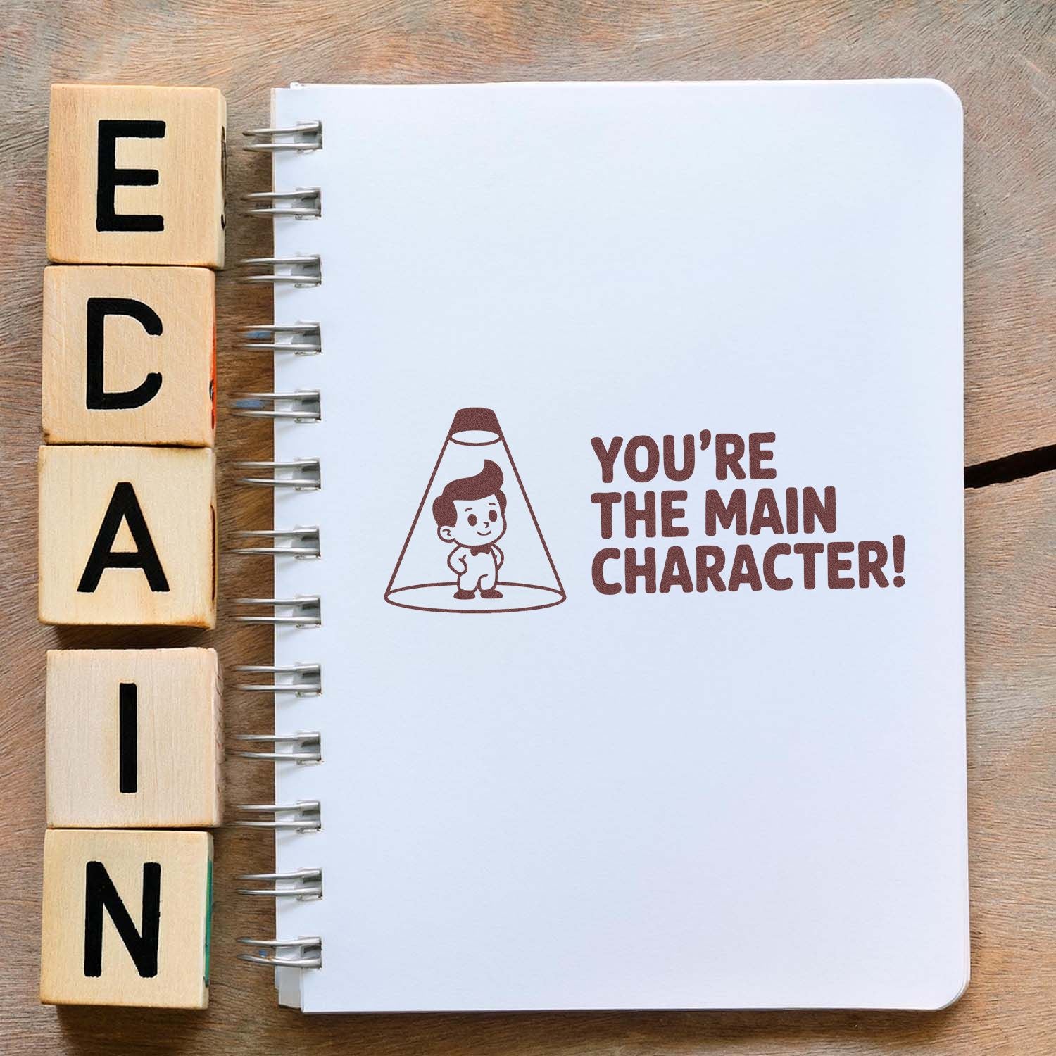 Wooden letter stamp blocks beside a white spiral notebook with a brown seal stamp that says Youre the main character!