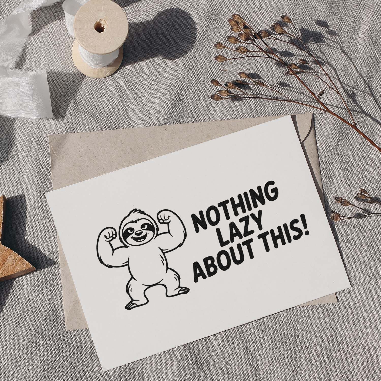 White stamp card with black flexing sloth and Nothing Lazy About This! on beige envelope, beside ribbon spool and dried stems.