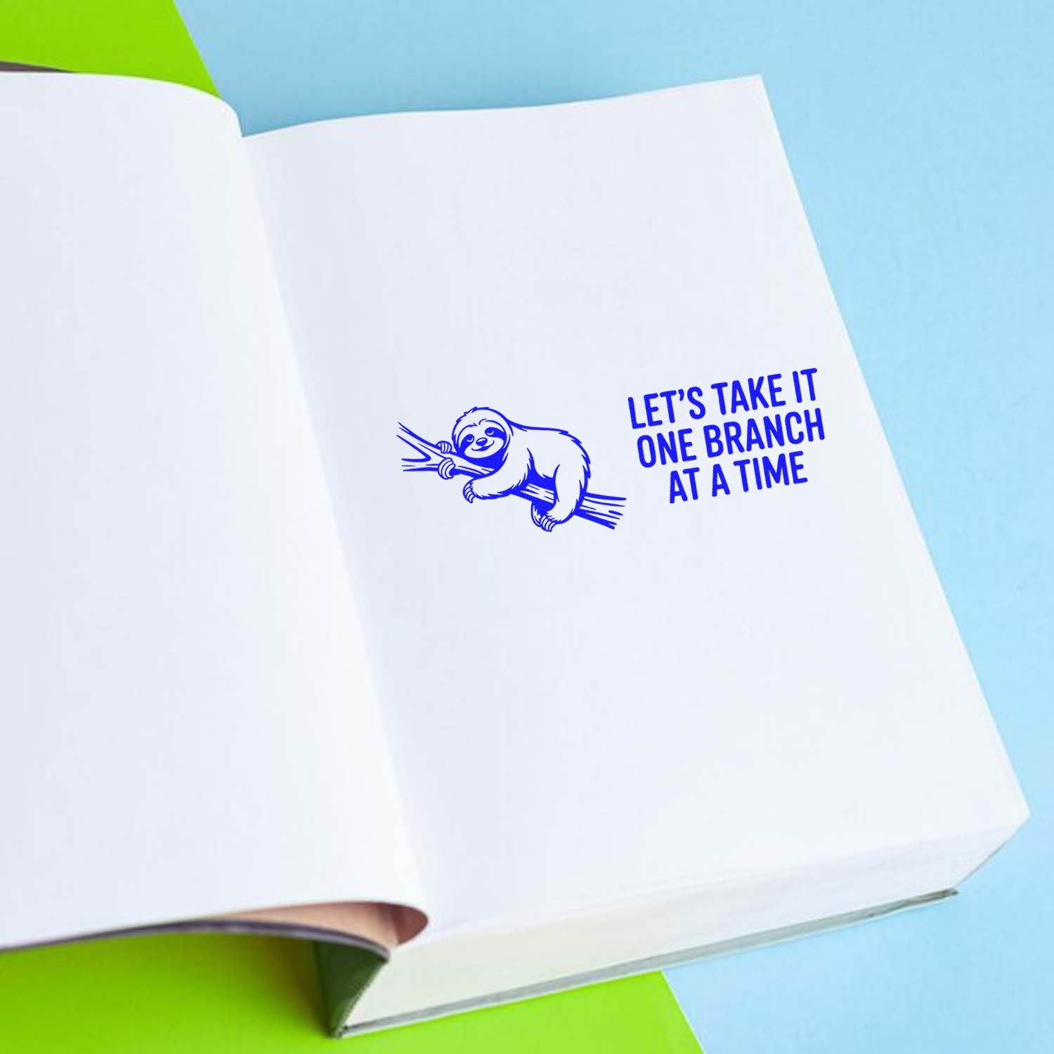 Blue sloth rubber stamp seal with Lets take it one branch at a time text on white paper against a light blue background.