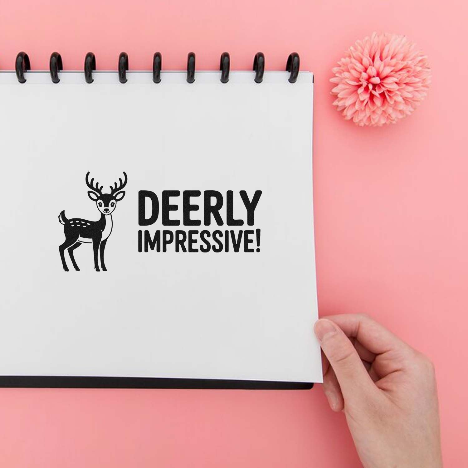 Black deer stamp and DEERLY IMPRESSIVE! seal on a white spiral notebook, set on a pink background with a pink flower.