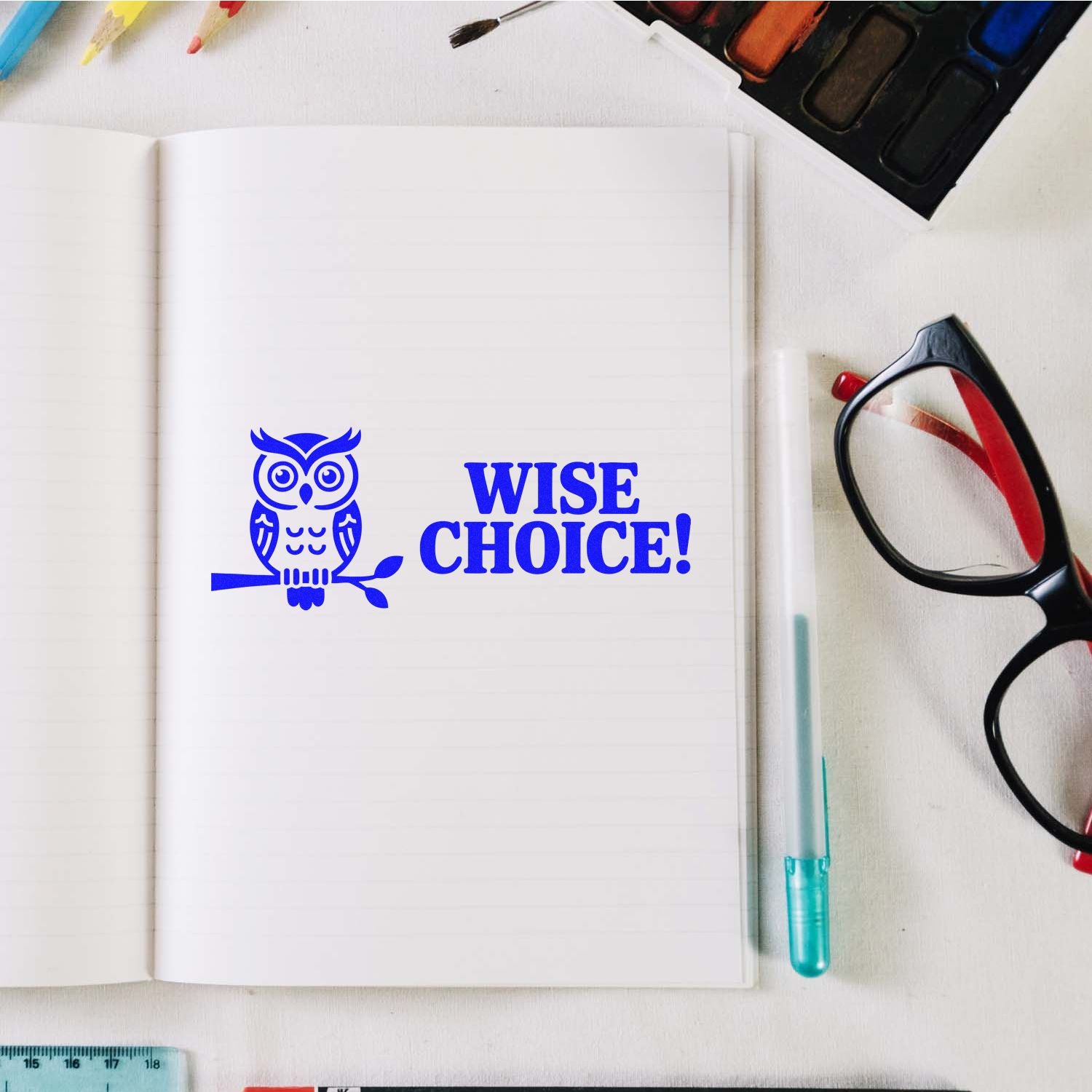 Blue owl stamp seal with WISE CHOICE! on an open notebook, beside pens, glasses, and paint palette.