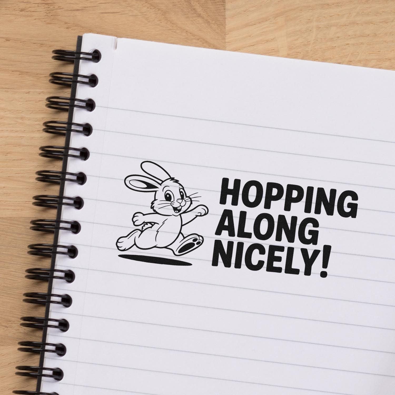 Black rabbit stamp and seal with HOPPING ALONG NICELY! on a white lined notebook, black spiral binding, wood background.