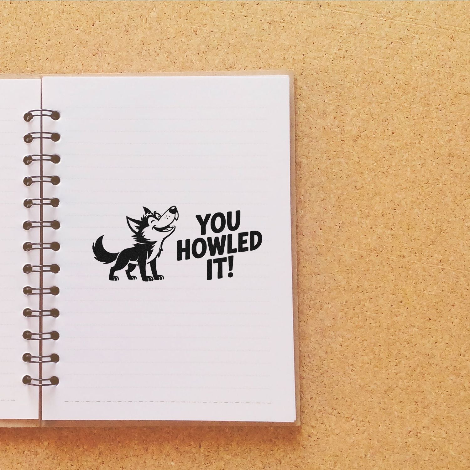 Black wolf stamp seal reading YOU HOWLED IT! on a white spiral notebook over a tan cork background.