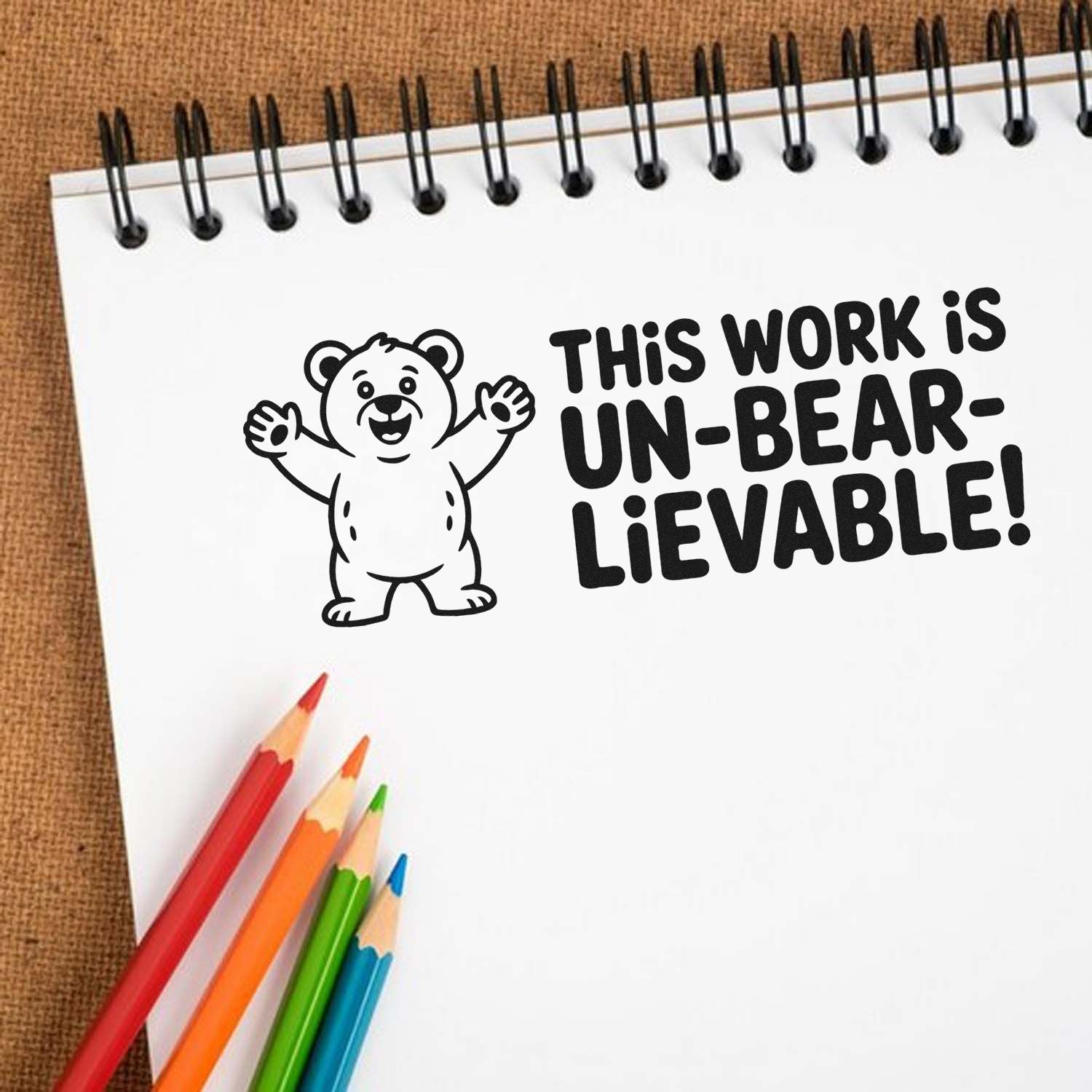 Black bear stamp with This work is un-bear-lievable! on a white spiral notebook, with red, orange, green and blue pencils.