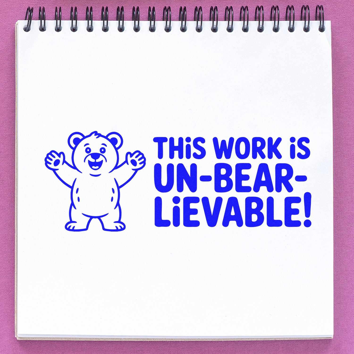 Blue bear stamp seal with This work is un-bear-lievable! on a white notepad against a pink background.