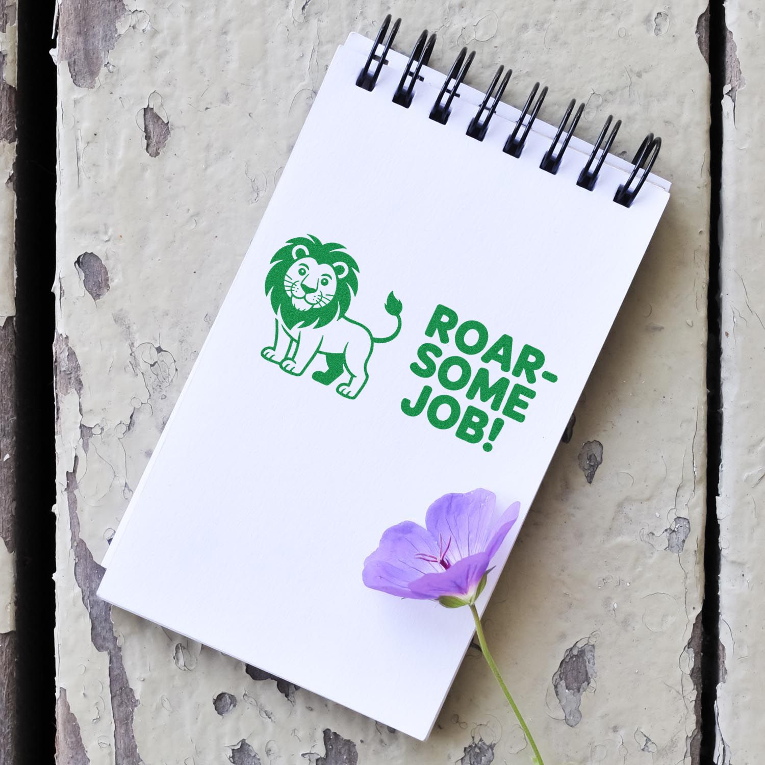 White spiral notebook with green lion stamp seal and ROAR-SOME JOB! text, accented by a purple flower on gray wood.