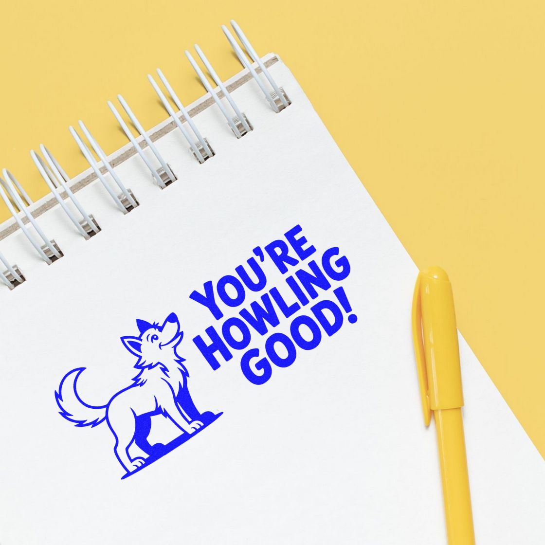 Blue dog stamp and seal reading Youre Howling Good! on a white spiral notebook, with a yellow pen on a yellow background.