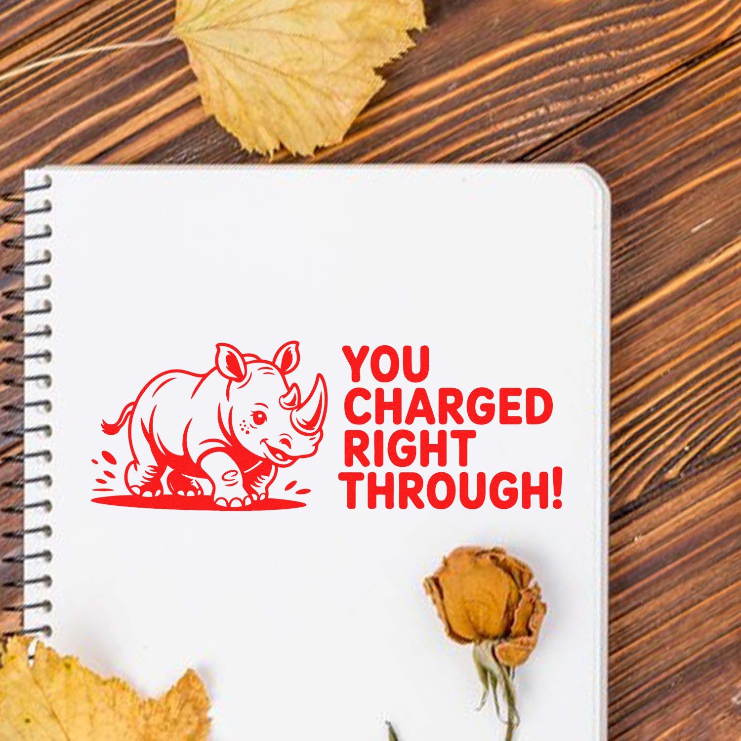 Red rhino self-inking stamp and seal with bold YOU CHARGED RIGHT THROUGH! on a white notebook page.