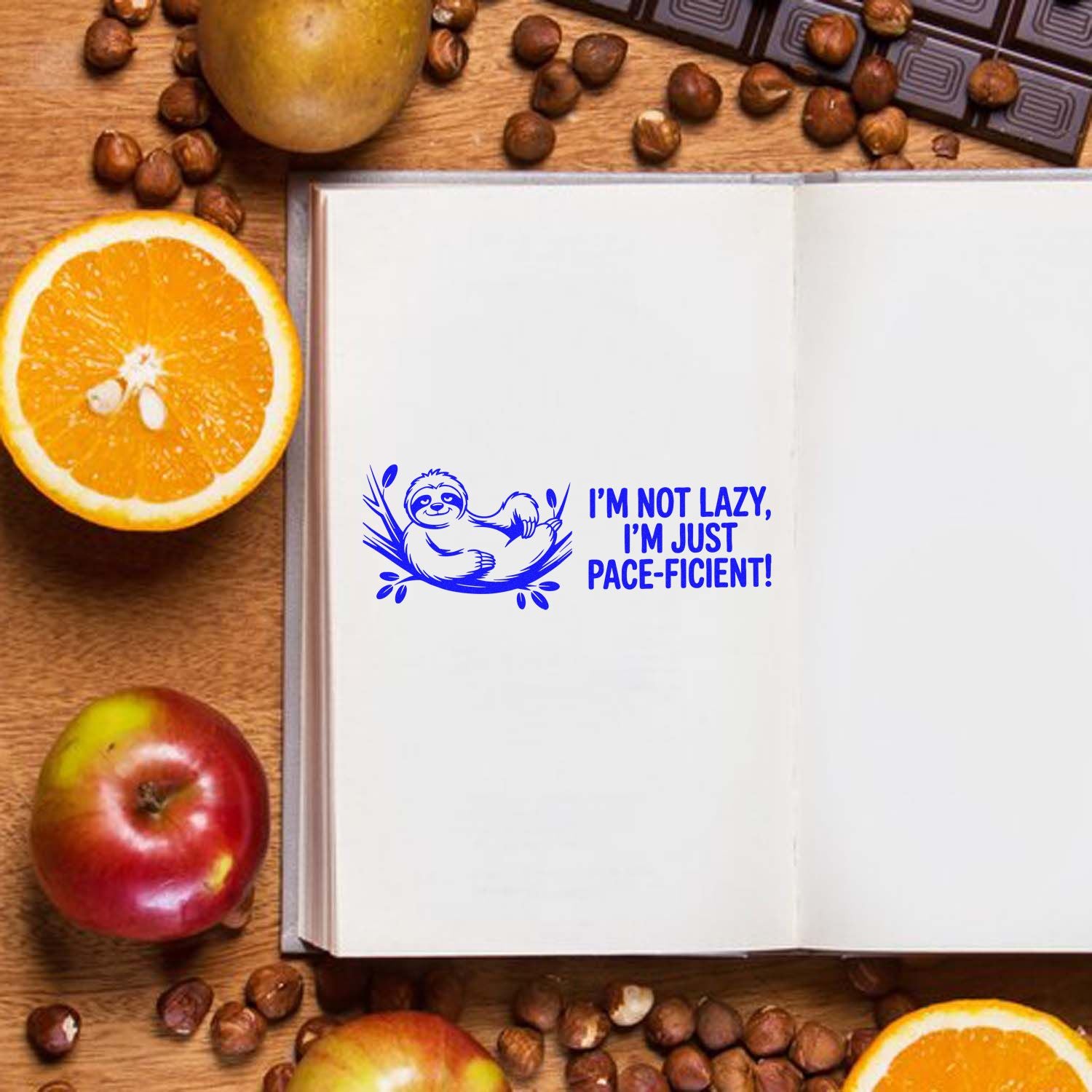 Blue sloth rubber stamp on open notebook, reading Im not lazy, Im just pace-ficient!, with oranges, apple and coffee beans.