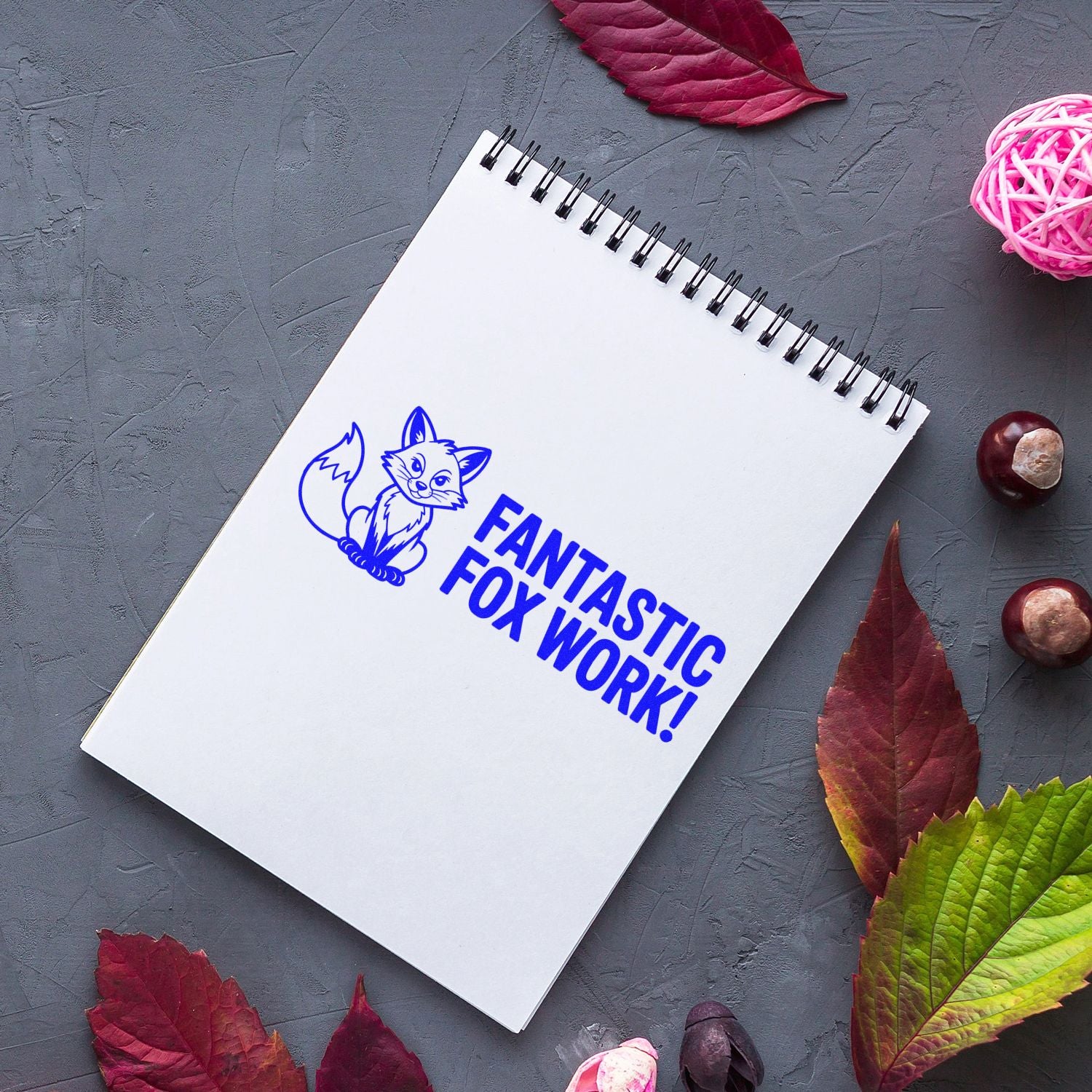 Blue custom stamp seal with fox logo and FANTASTIC FOX WORK! on white spiral notebook, surrounded by autumn leaves.