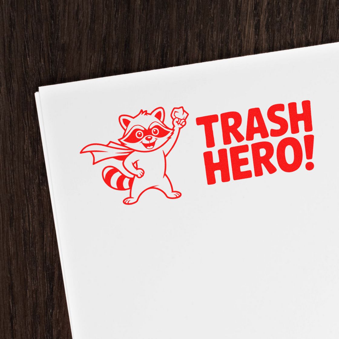 Red raccoon Trash Hero stamp and seal design on white paper, bold red lettering, dark wood background.