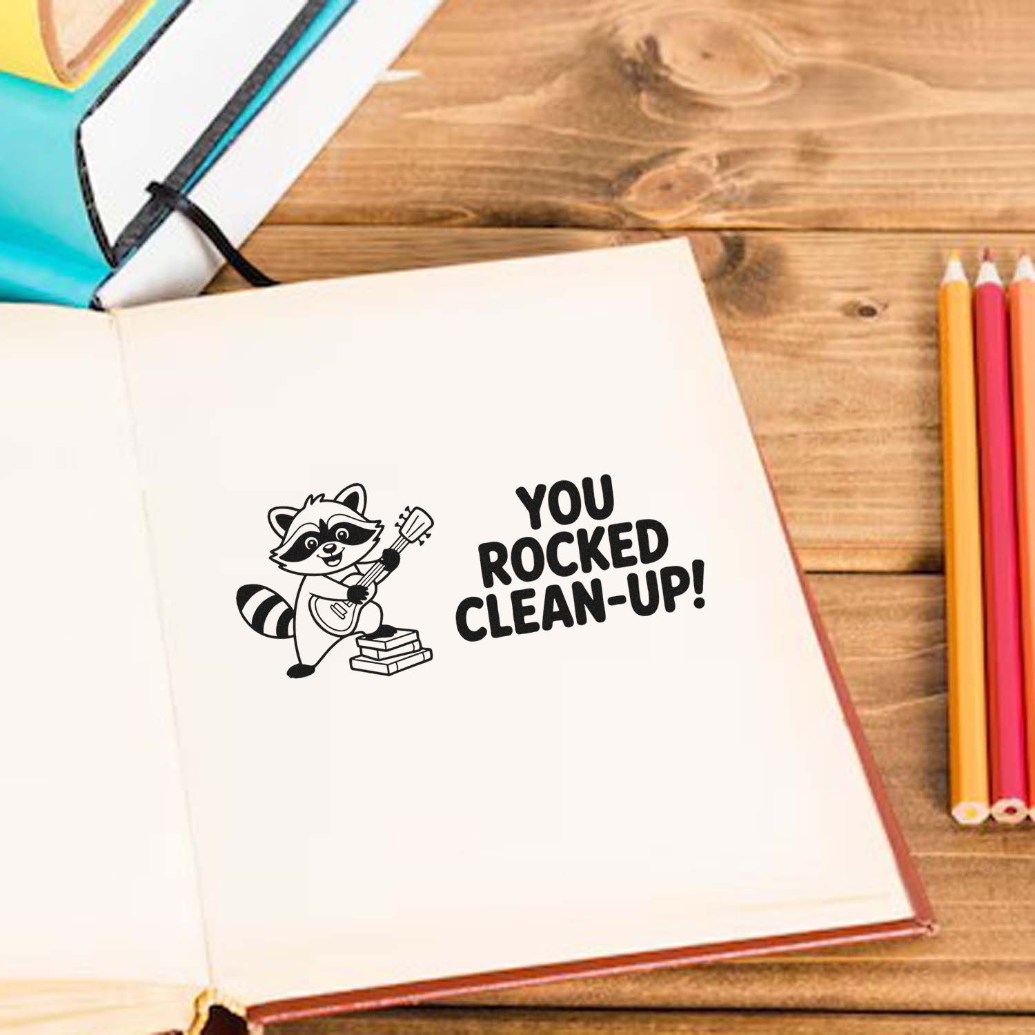 Black stamp and seal with raccoon guitar design and You rocked clean-up! on white notebook, beside colorful pencils.