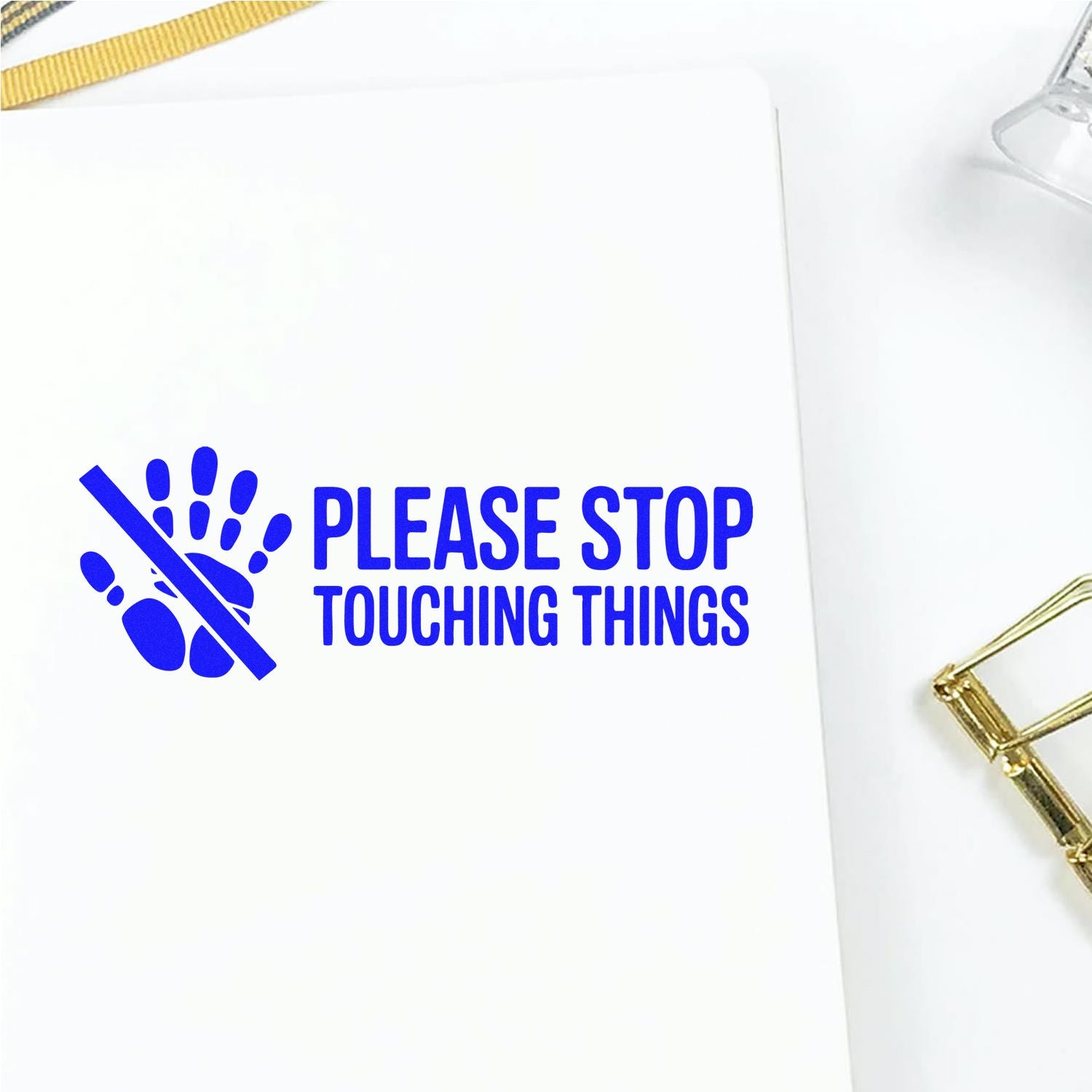 Blue self-inking stamp reading Please Stop Touching Things with a hand icon on white paper, accented by gold clip and ribbon.