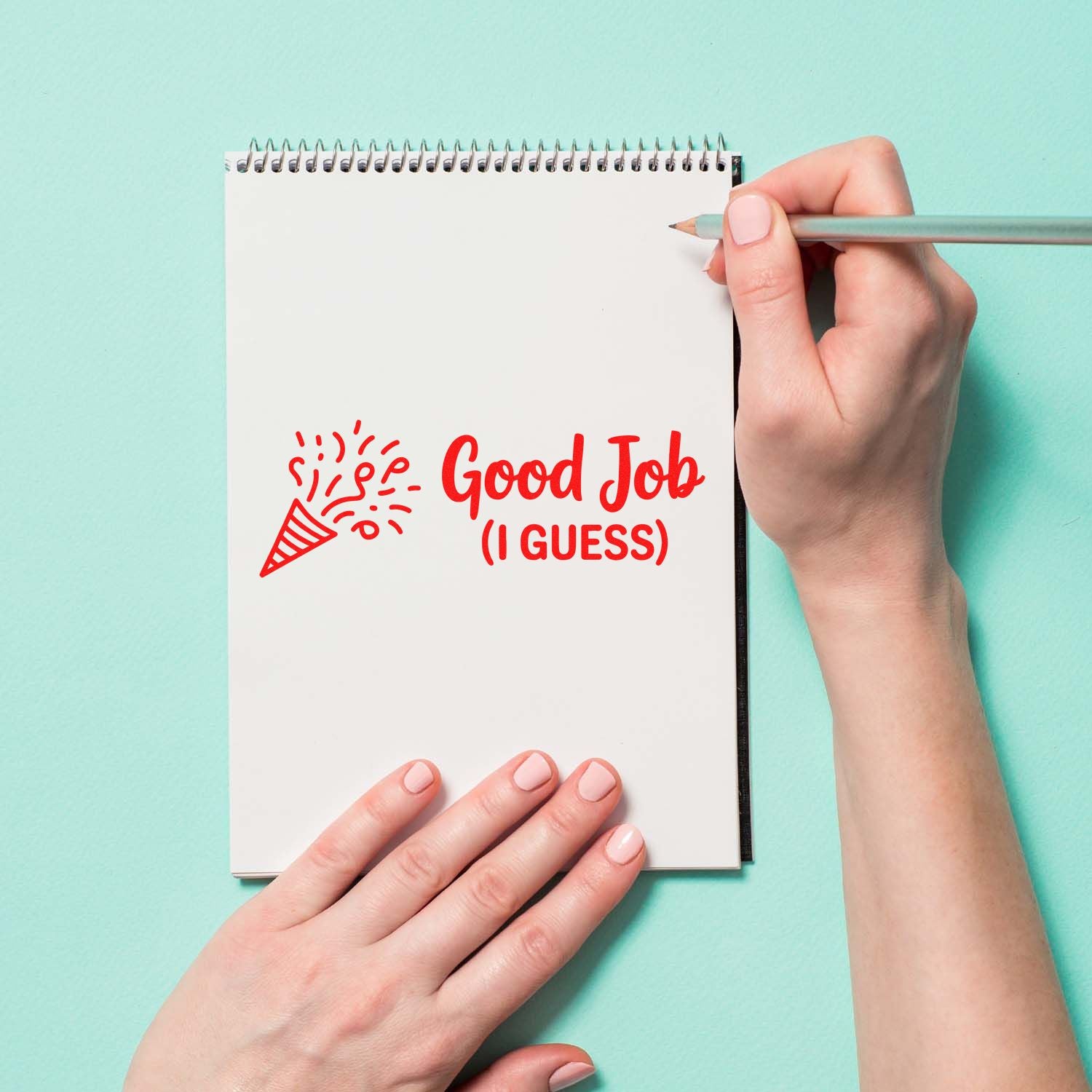 Red self-inking stamp reading Good Job (I GUESS) with party popper, on a white spiral notebook against a mint background.