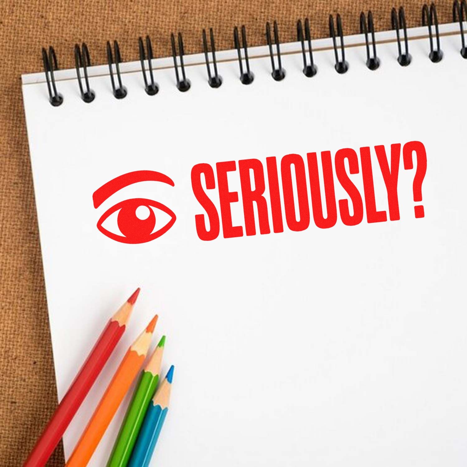 Red Seriously? stamp with eye icon on a white spiral notepad, beside colorful pencils.