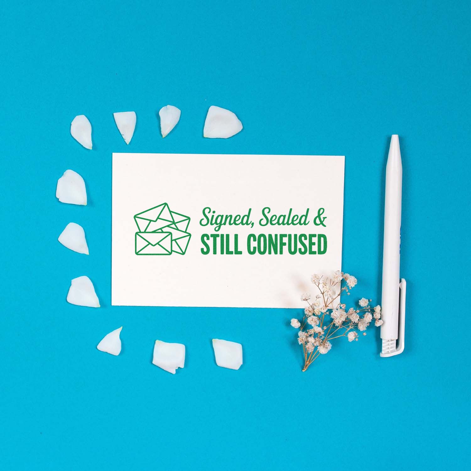 Green envelope stamp design on white card, with white wax seal pieces, white pen, and dried flowers on bright blue background.