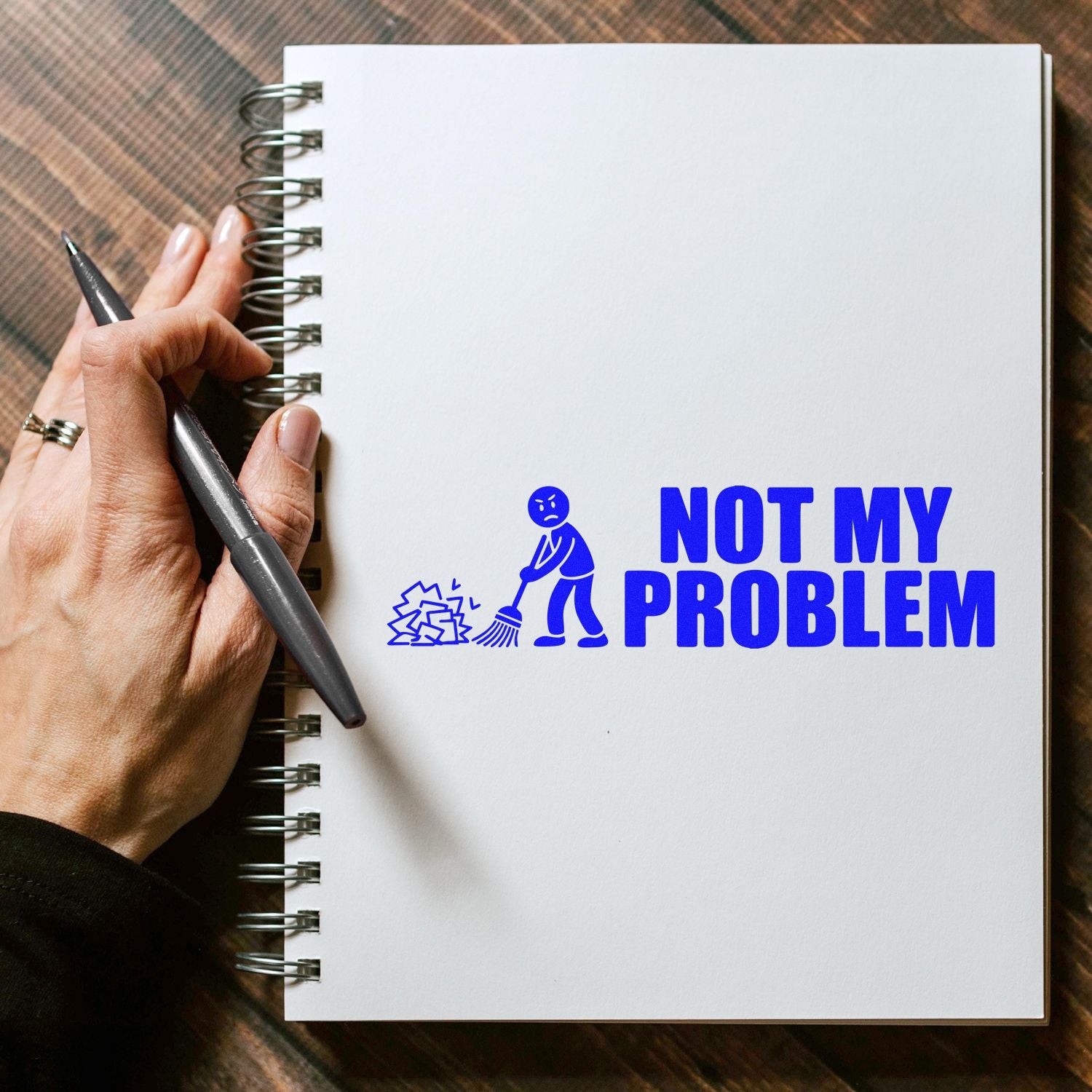 Blue Not My Problem stamp seal on a white spiral notebook, with cleanup icon and hand holding a pen.