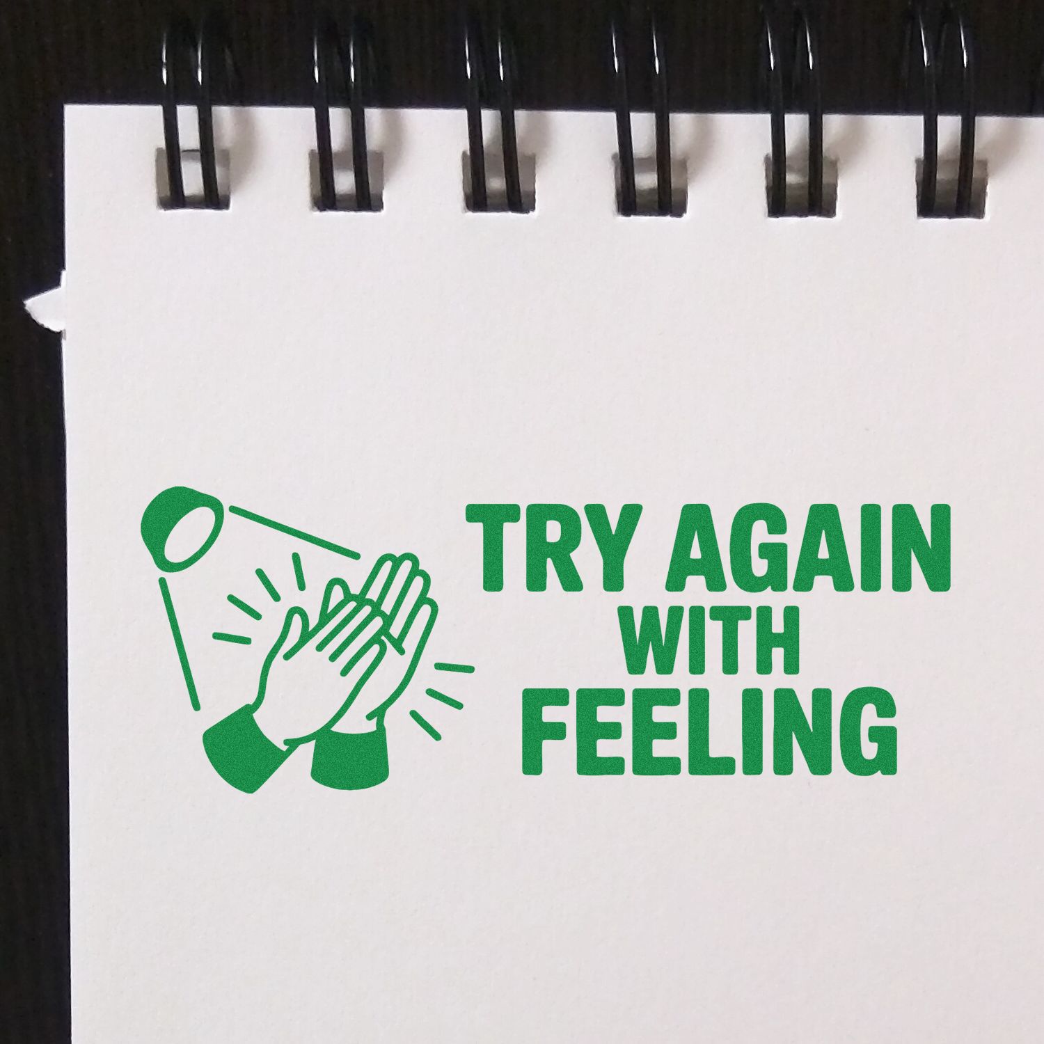Green rubber stamp design with clapping hands icon and bold text TRY AGAIN WITH FEELING on a white notepad.