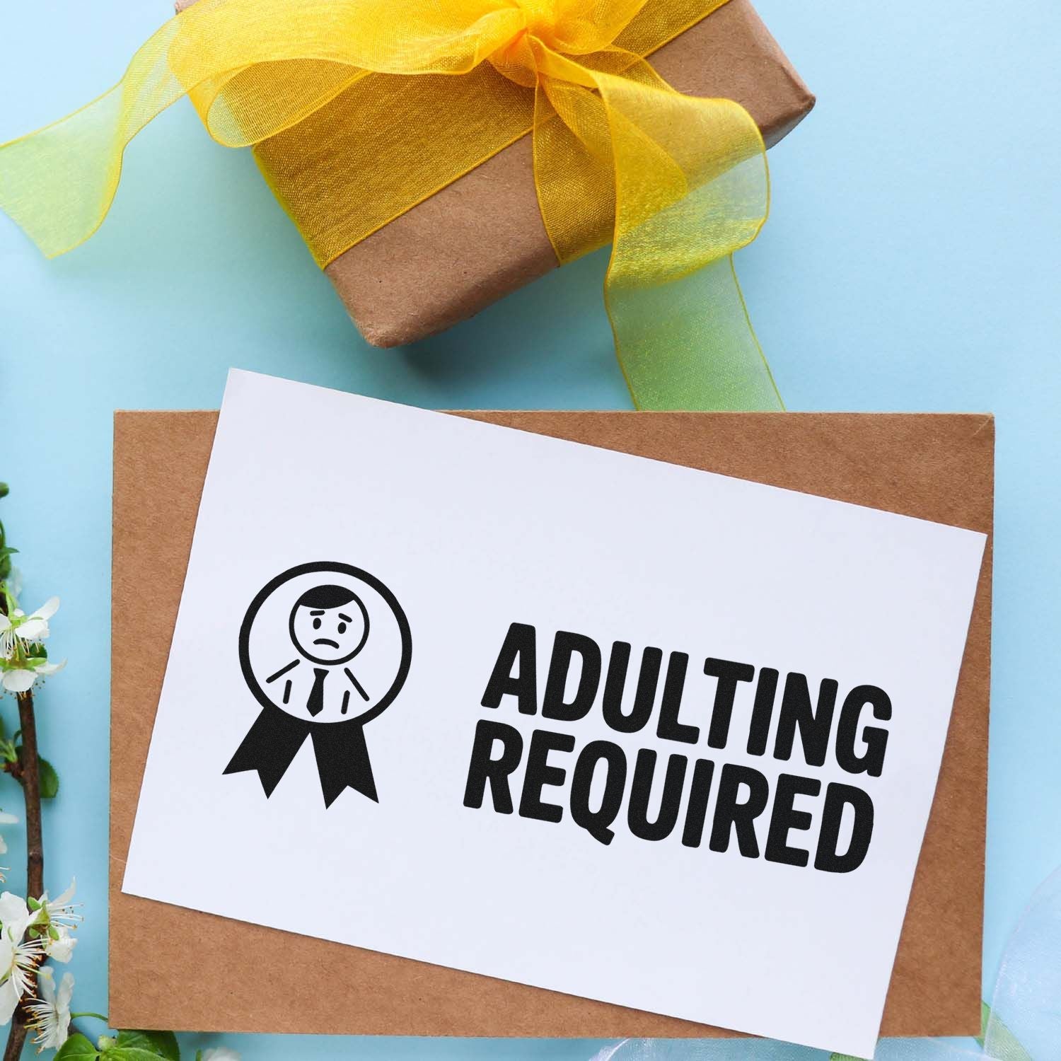 Brown kraft gift package with yellow ribbon, featuring a white stamp card reading ADULTING REQUIRED with black seal icon.