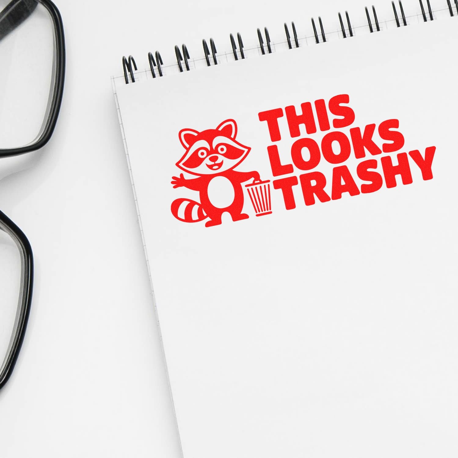 Red custom stamp design with raccoon and trash can on white spiral notebook reading THIS LOOKS TRASHY, beside black eyeglasses.