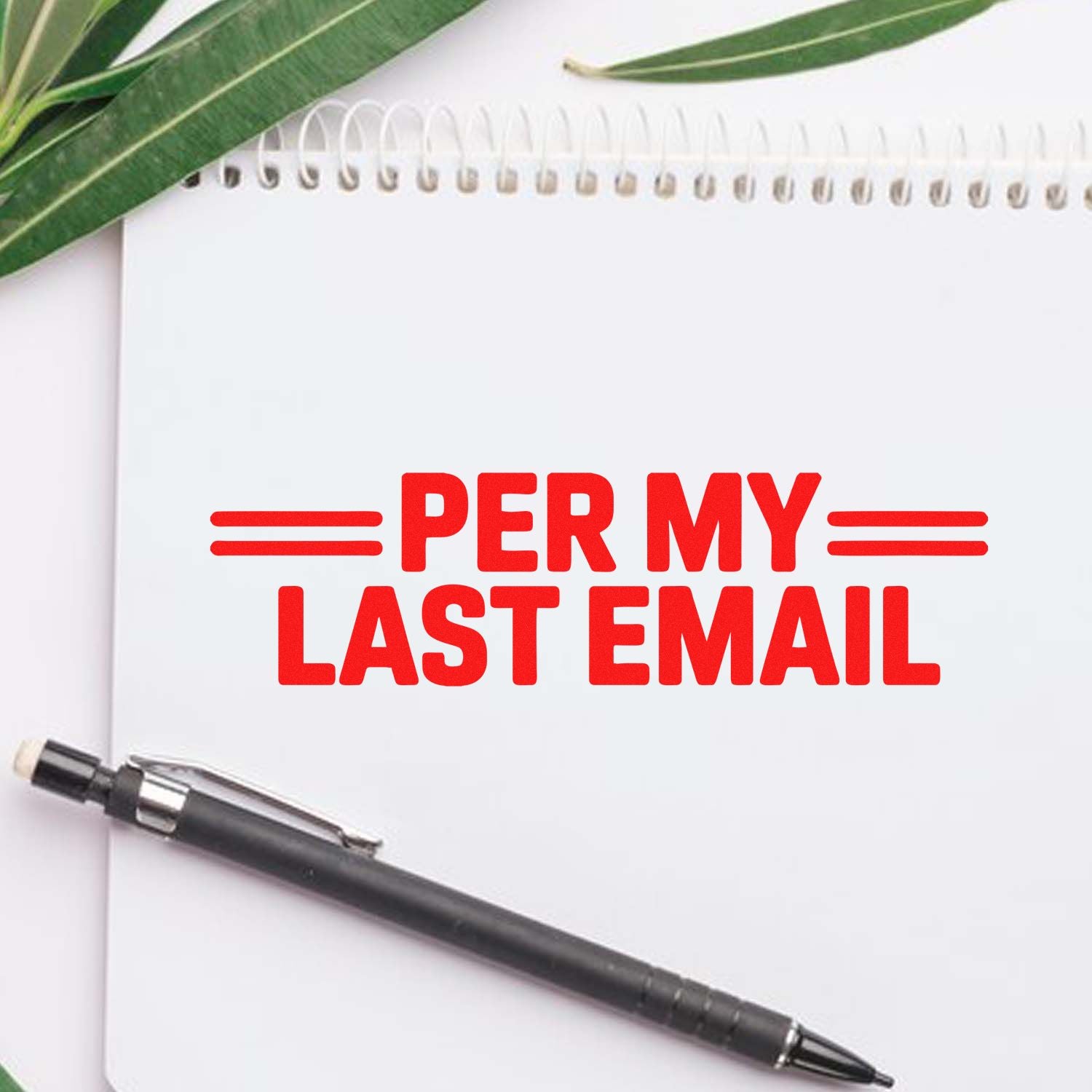 Red PER MY LAST EMAIL stamp on a white spiral notebook, with a black pen and green leaves.